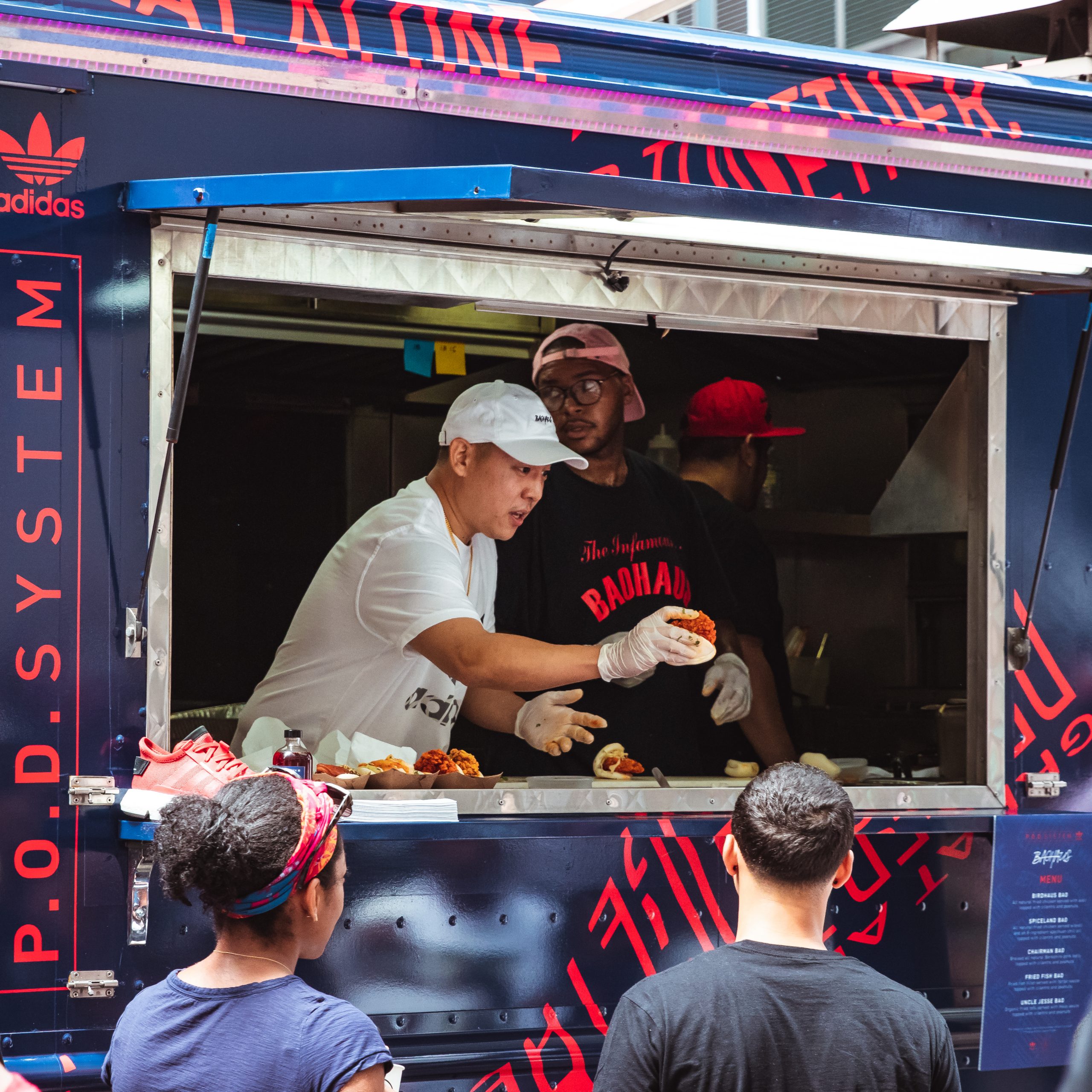 Two food truck workers serve customers at a blue truck with red text; people wait in line.