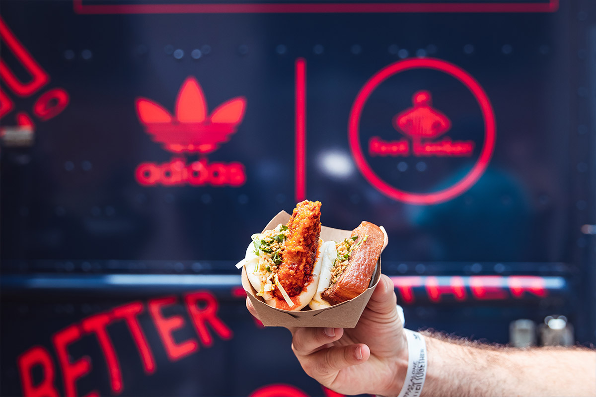 Hand holding tray with two fried chicken sandwiches in front of a blue Adidas-branded food truck.