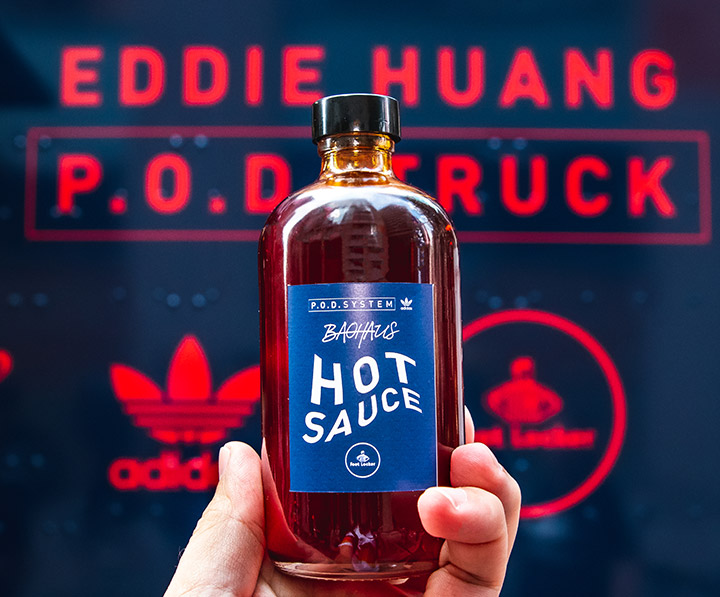 Hand holding blue-label hot sauce in front of Eddie Huang P.O.D. Truck with Adidas logos behind.