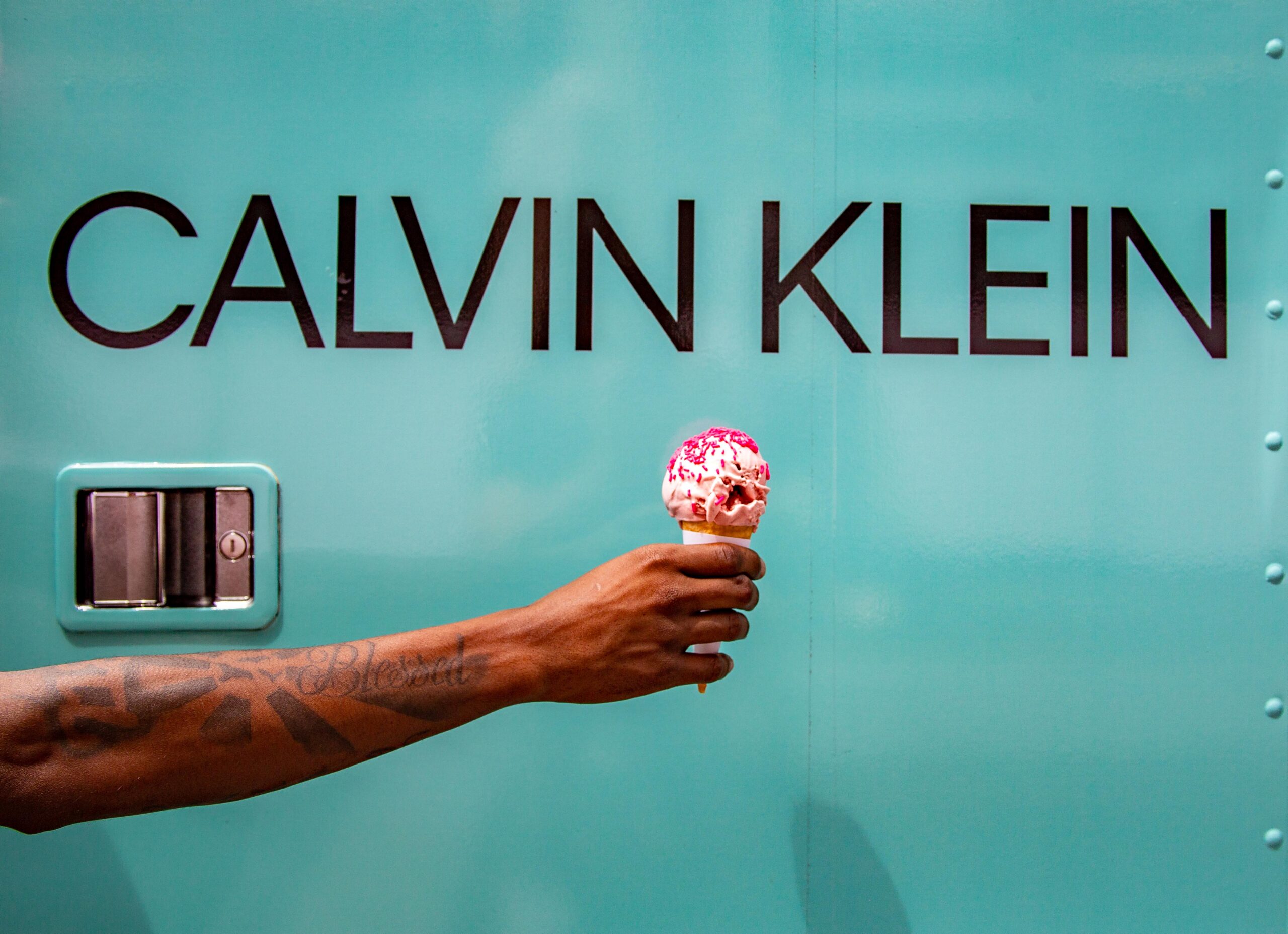 Tattooed arm holding pink-sprinkled ice cream; CALVIN KLEIN logo and truck reflected nearby.