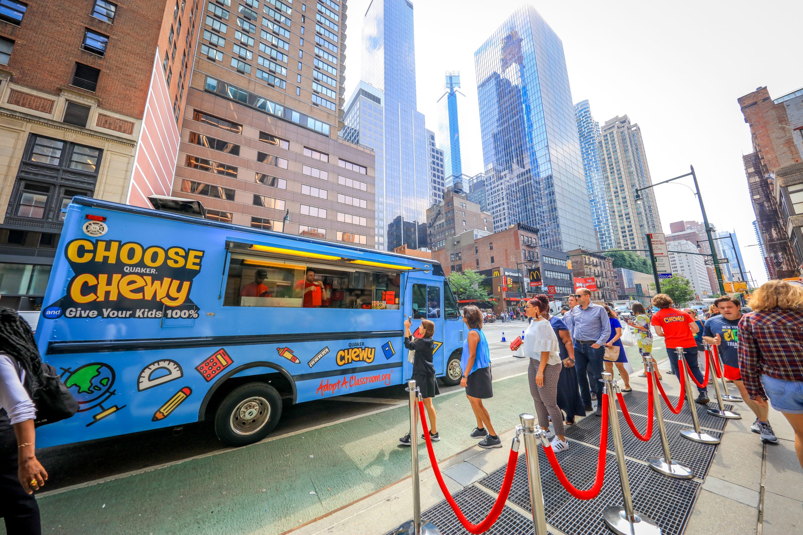 Blue food truck with Choose Chewy branding parked on a city street; people line up nearby.