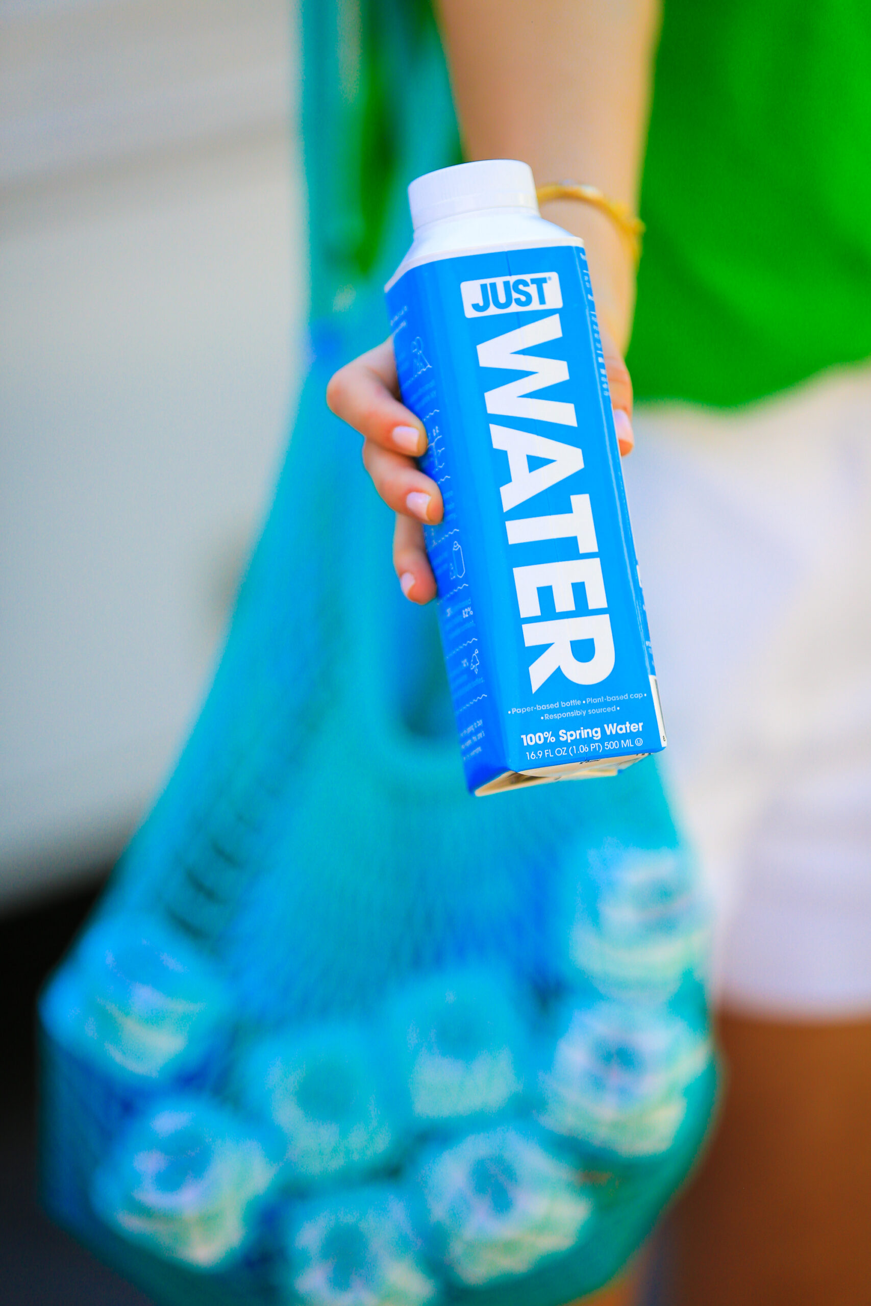Person in green top and white shorts holds a blue JUST Water carton near a bag of more cartons.