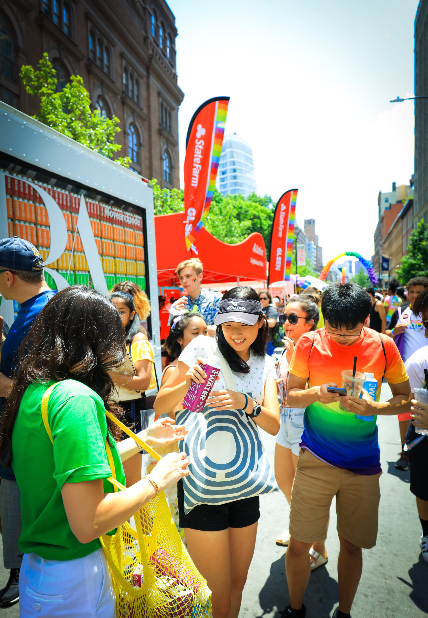 People laughing and drinking at an outdoor Pride parade with rainbow flags and banners nearby.