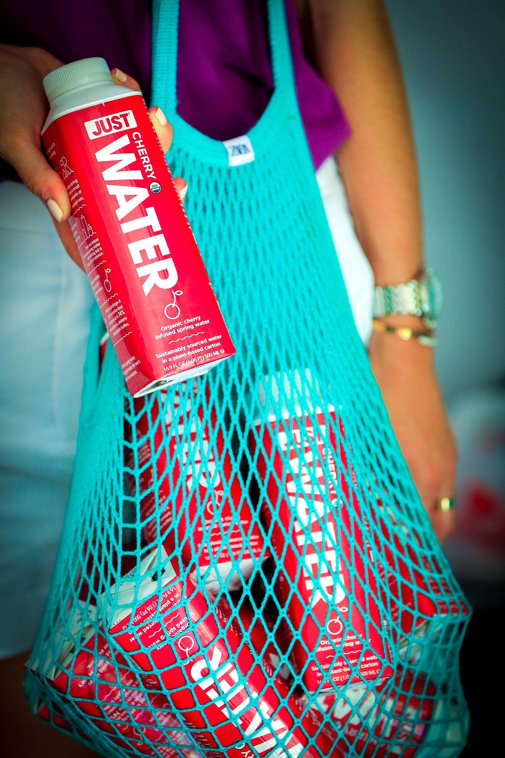 Person holding a turquoise mesh bag of red JUST WATER cartons, wearing purple top and white shorts.