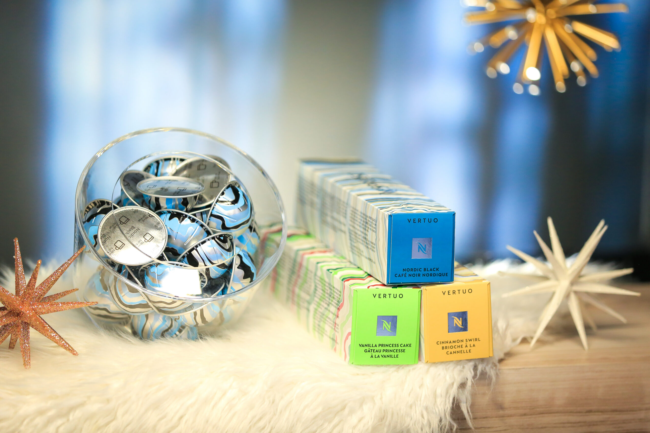 Glass bowl of Nespresso pods beside three Vertuo capsule box stacks on white surface with stars.