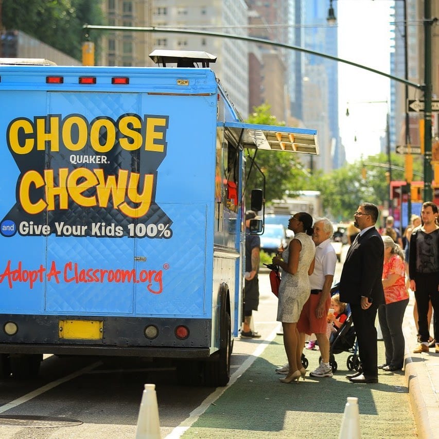 People line up at a blue Quaker Chewy food truck promoting Adopt A Classroom.org on a city street.