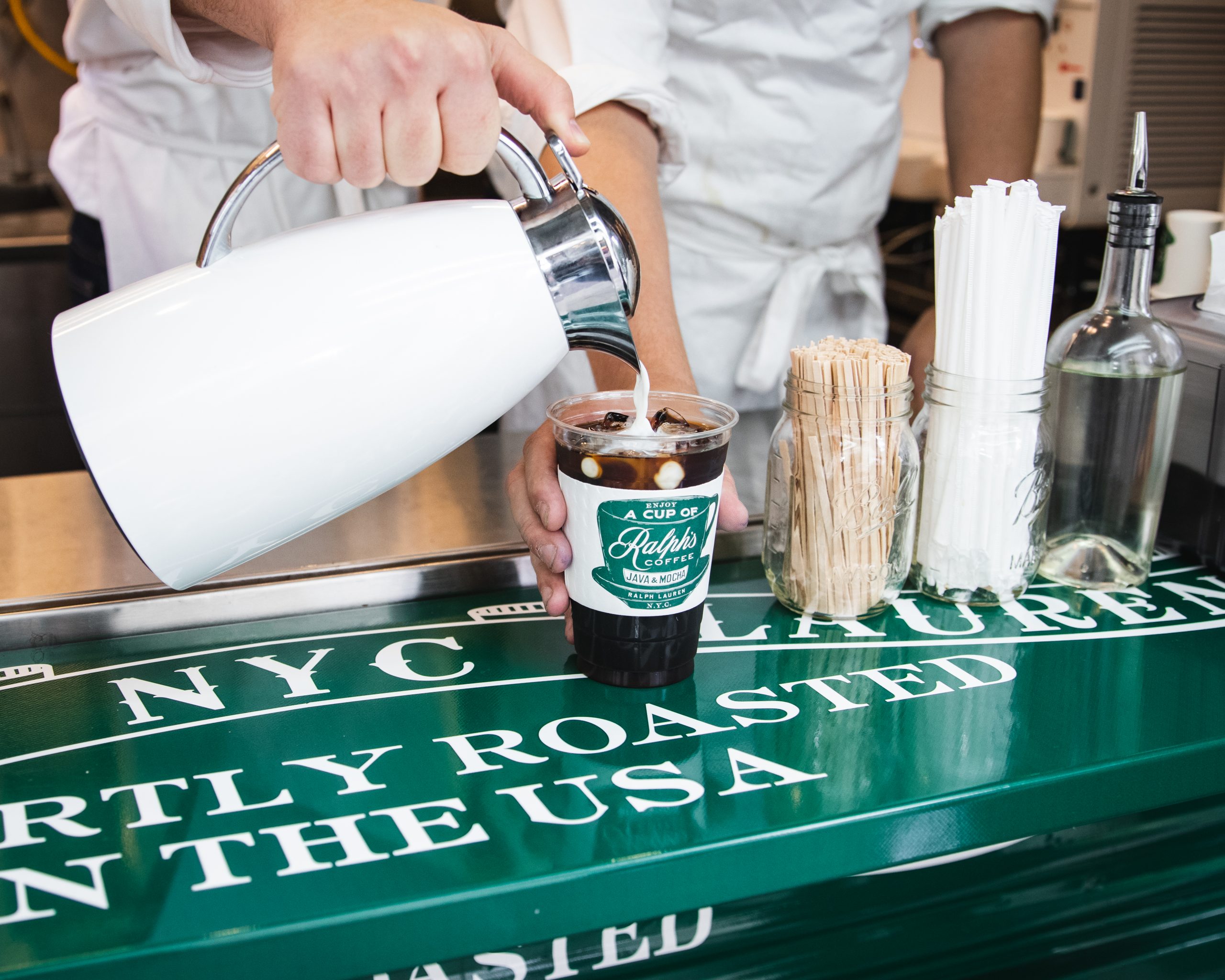Person pours from white pitcher into iced coffee at NYC LAUREL counter with straws and a bottle.