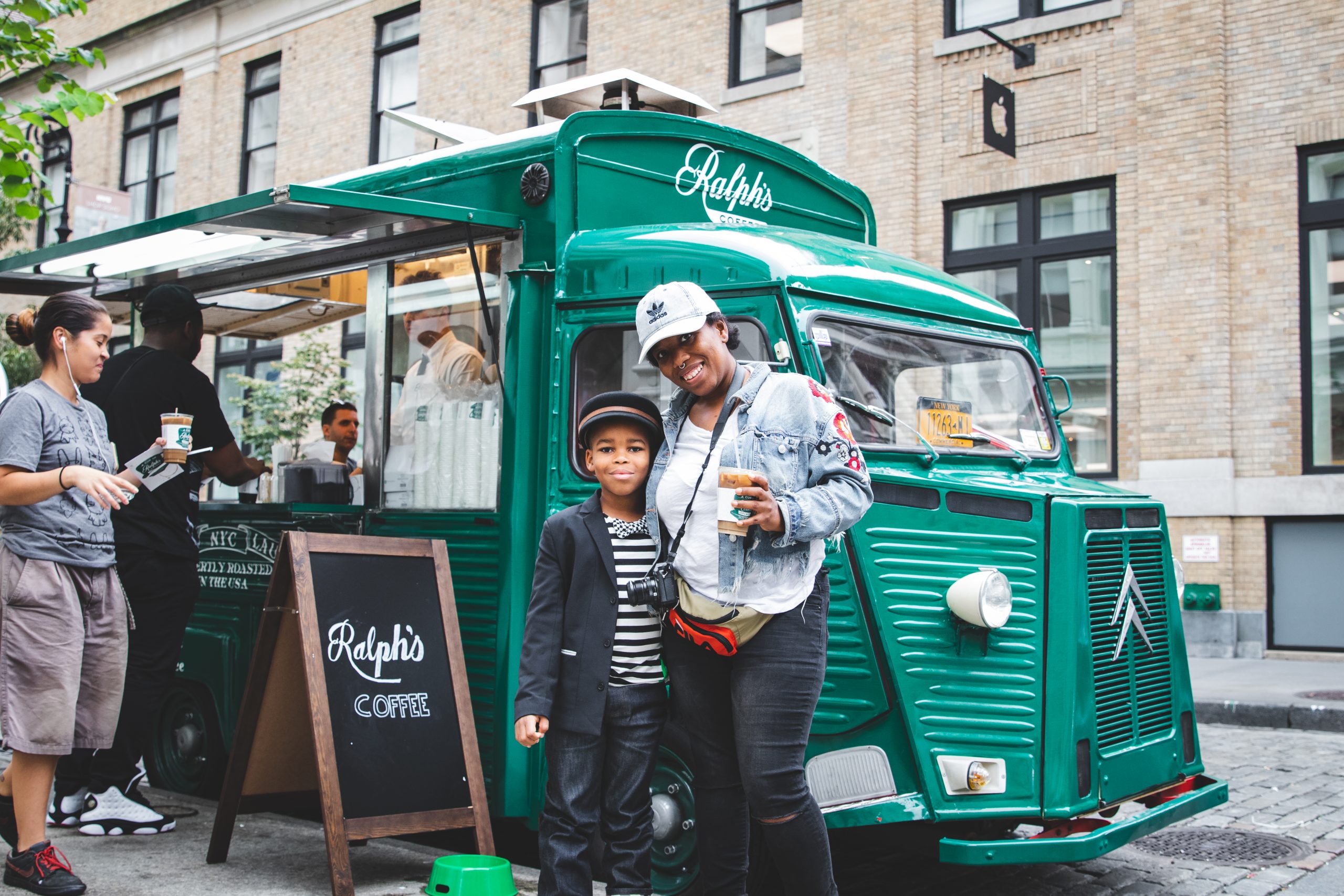 A woman and child smile by a green Ralph's Coffee truck on a city street with people nearby.