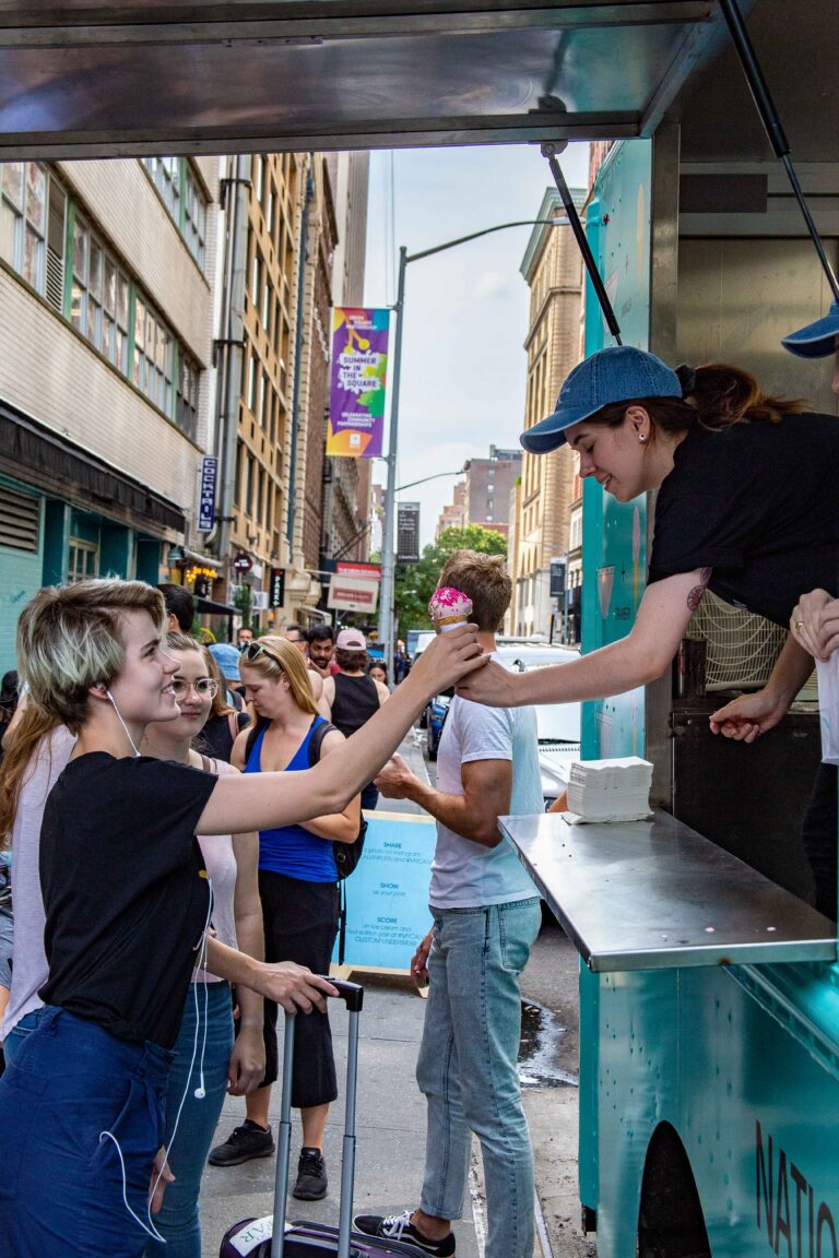 A woman gets an ice cream cone from a worker at a truck on a busy city street.