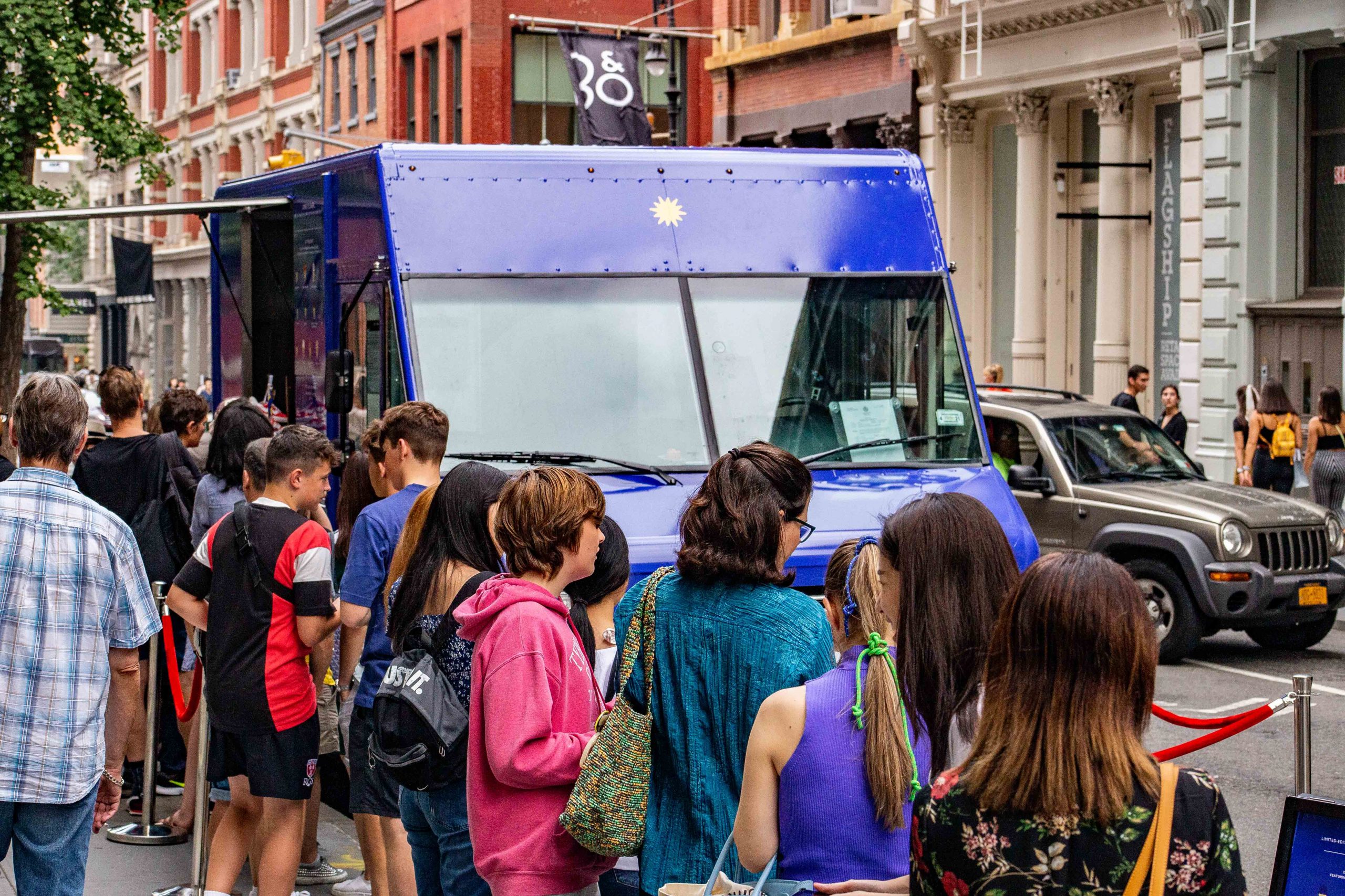 People line up outside a blue food truck on a city sidewalk as a gray SUV passes by.