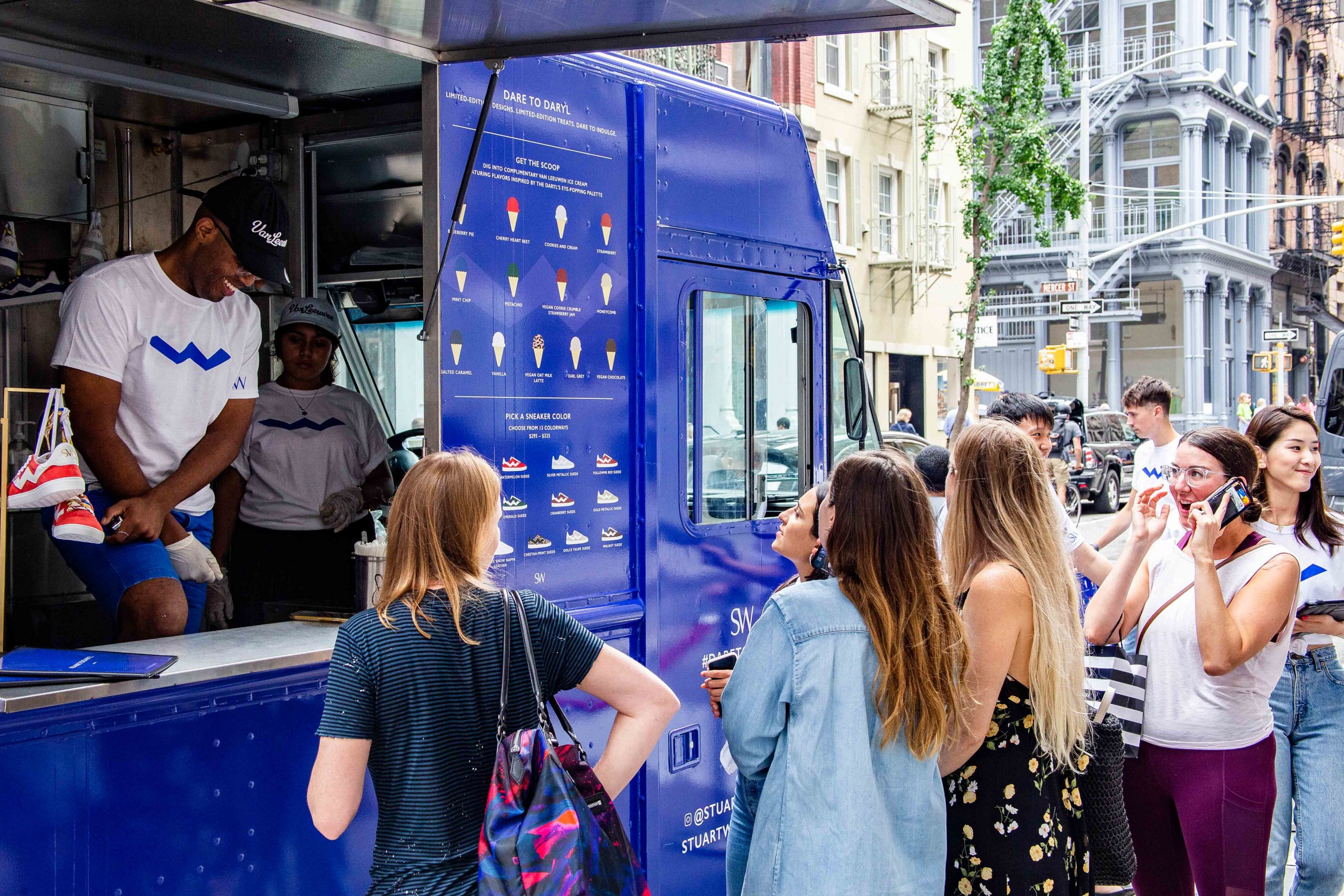 People line up at a blue food truck on a city street as a staff member serves a customer.