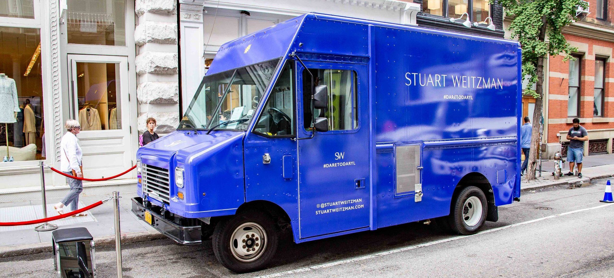 Stuart Weitzman blue delivery truck parked outside a boutique, with people and red ropes nearby.
