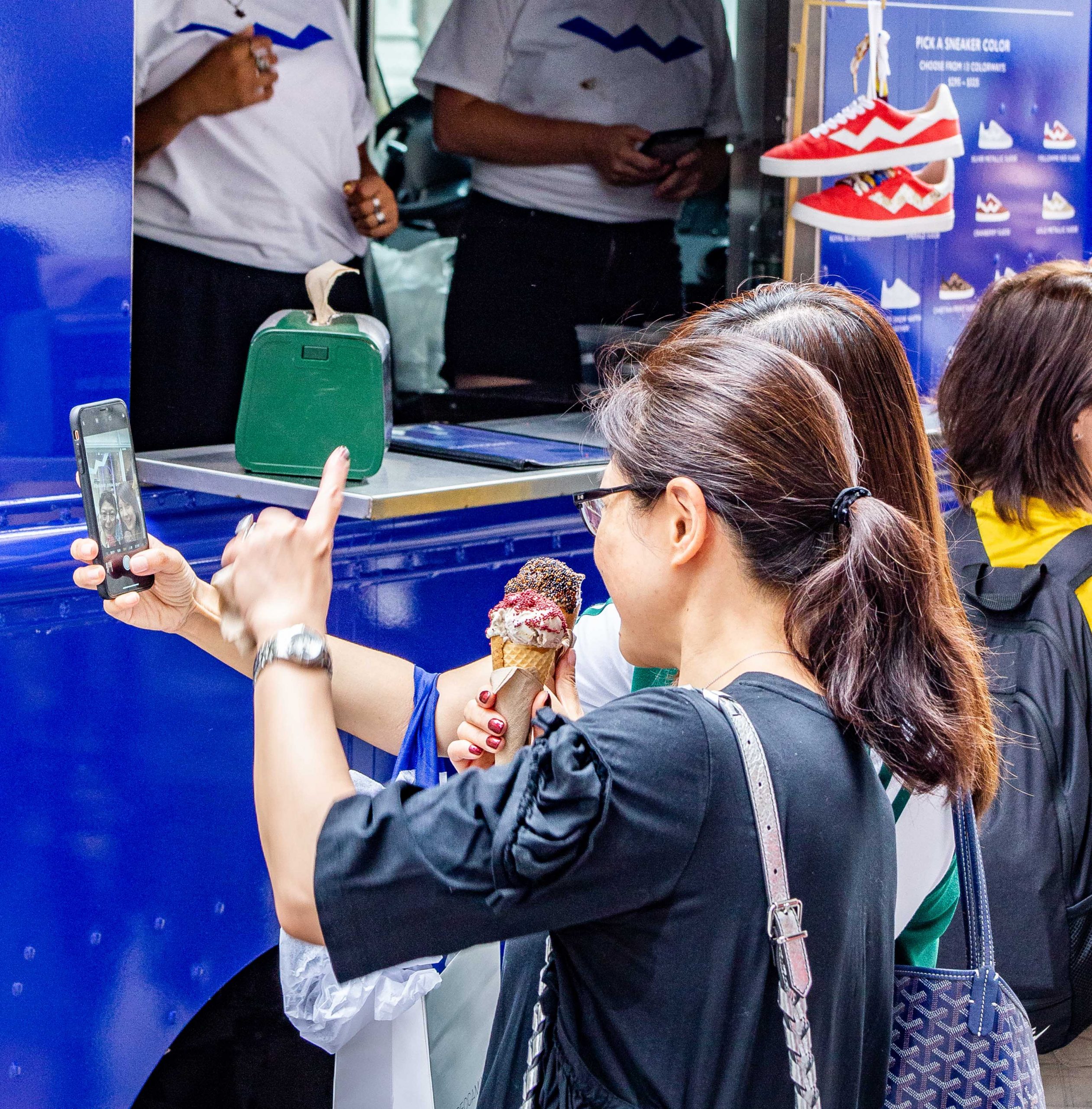 Two women by a blue food truck; one eats ice cream, the other takes a selfie. Shoes are on display.