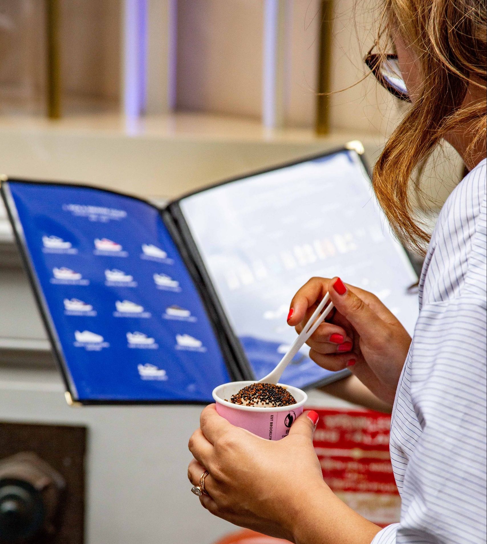 Woman with painted nails stirs ice cream topped with chocolate sprinkles while reading a blue menu.