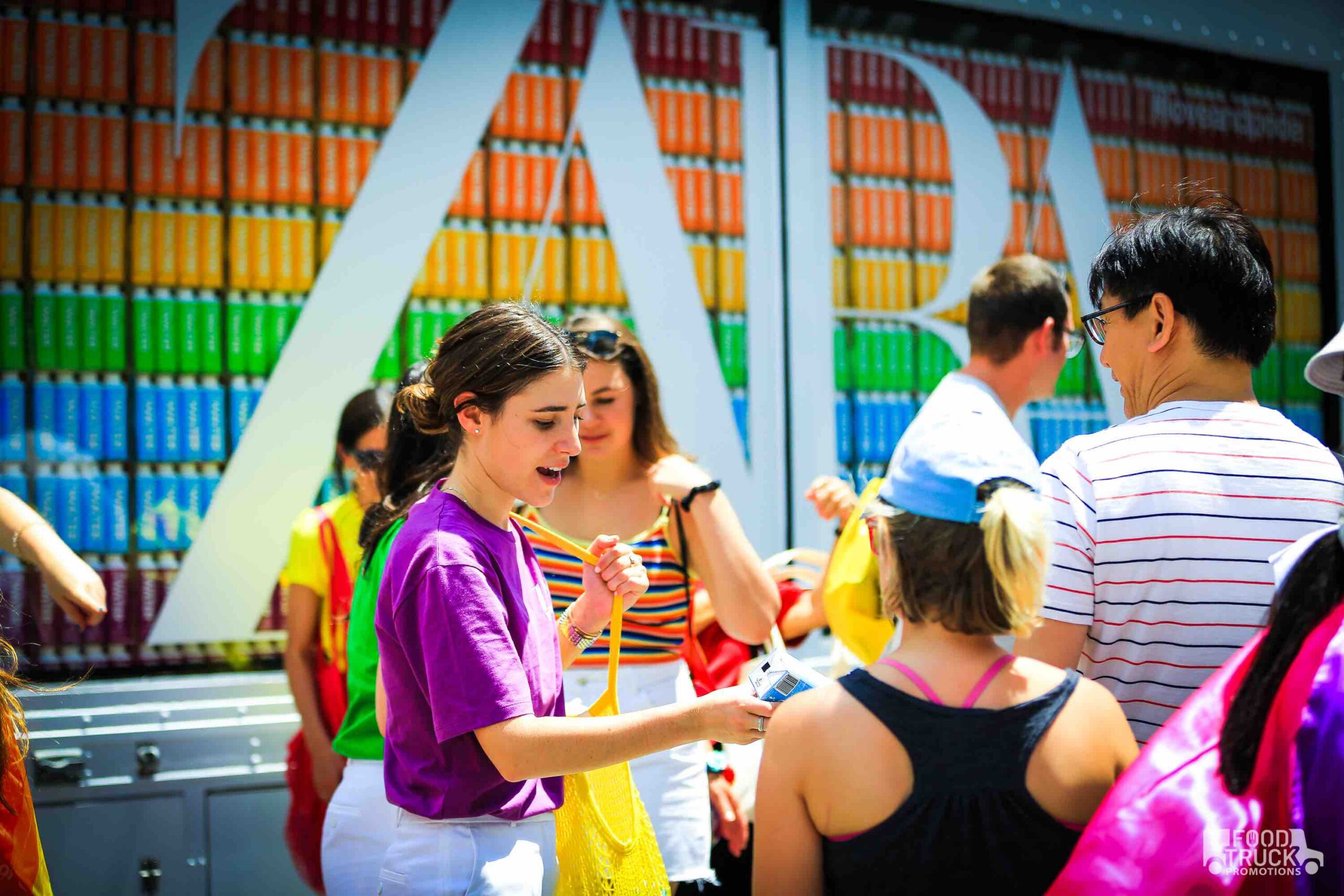 Woman in purple shirt gives items to group near colorful cans and large white ZARA letters.
