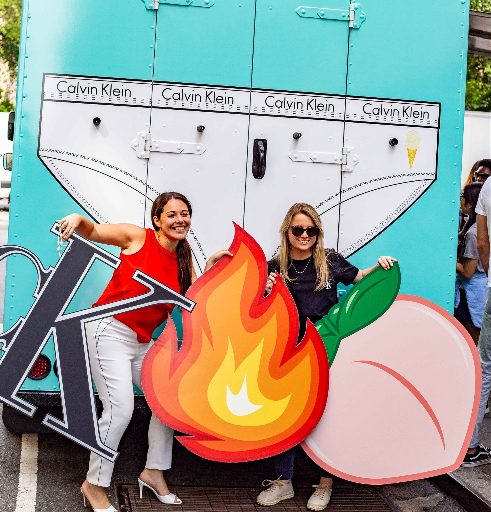 Two women smile in front of a Calvin Klein truck, holding large cutouts of a K, fire, and peach.