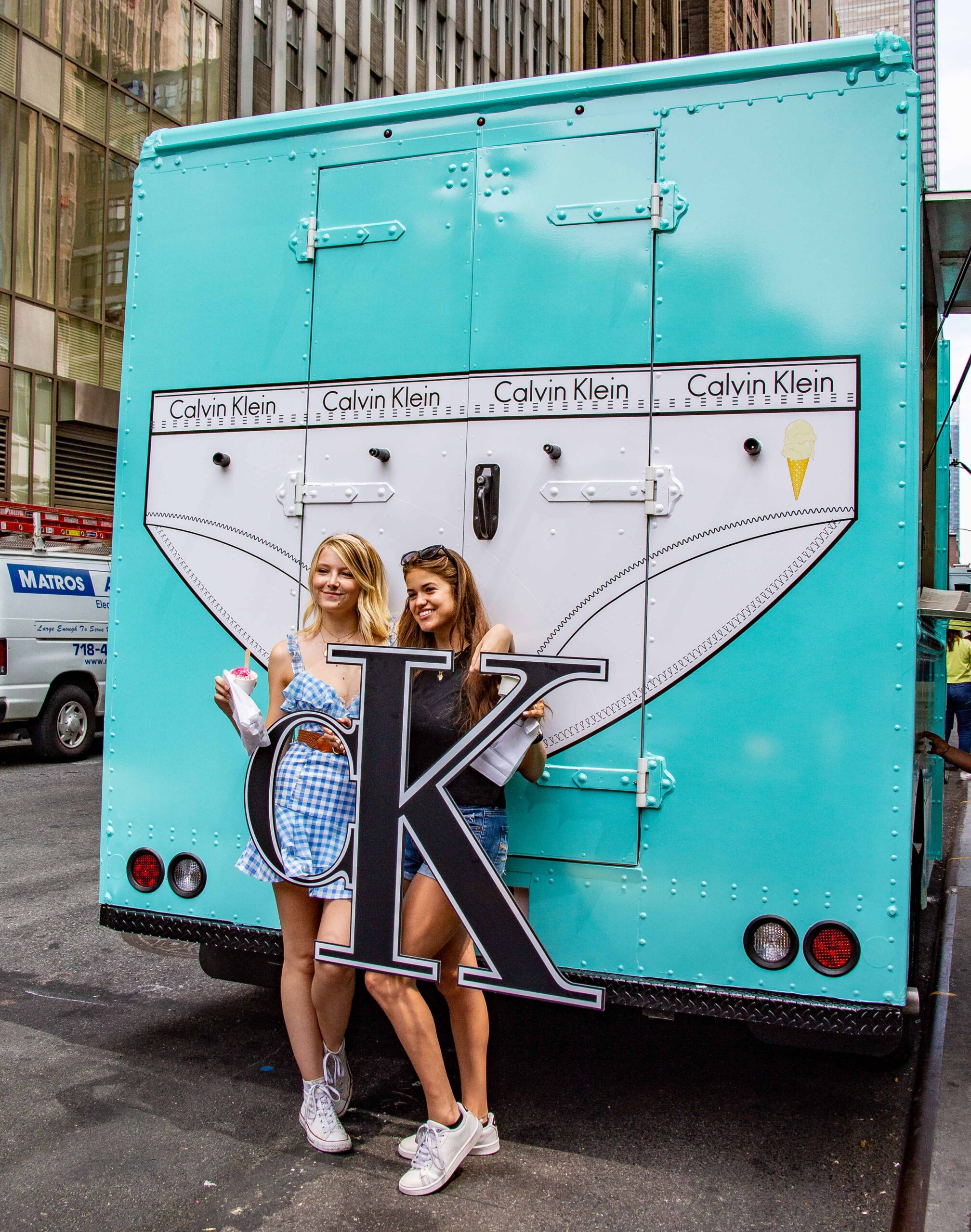 Two women hold a CK sign and ice cream cones in front of a Calvin Klein truck on a city street.