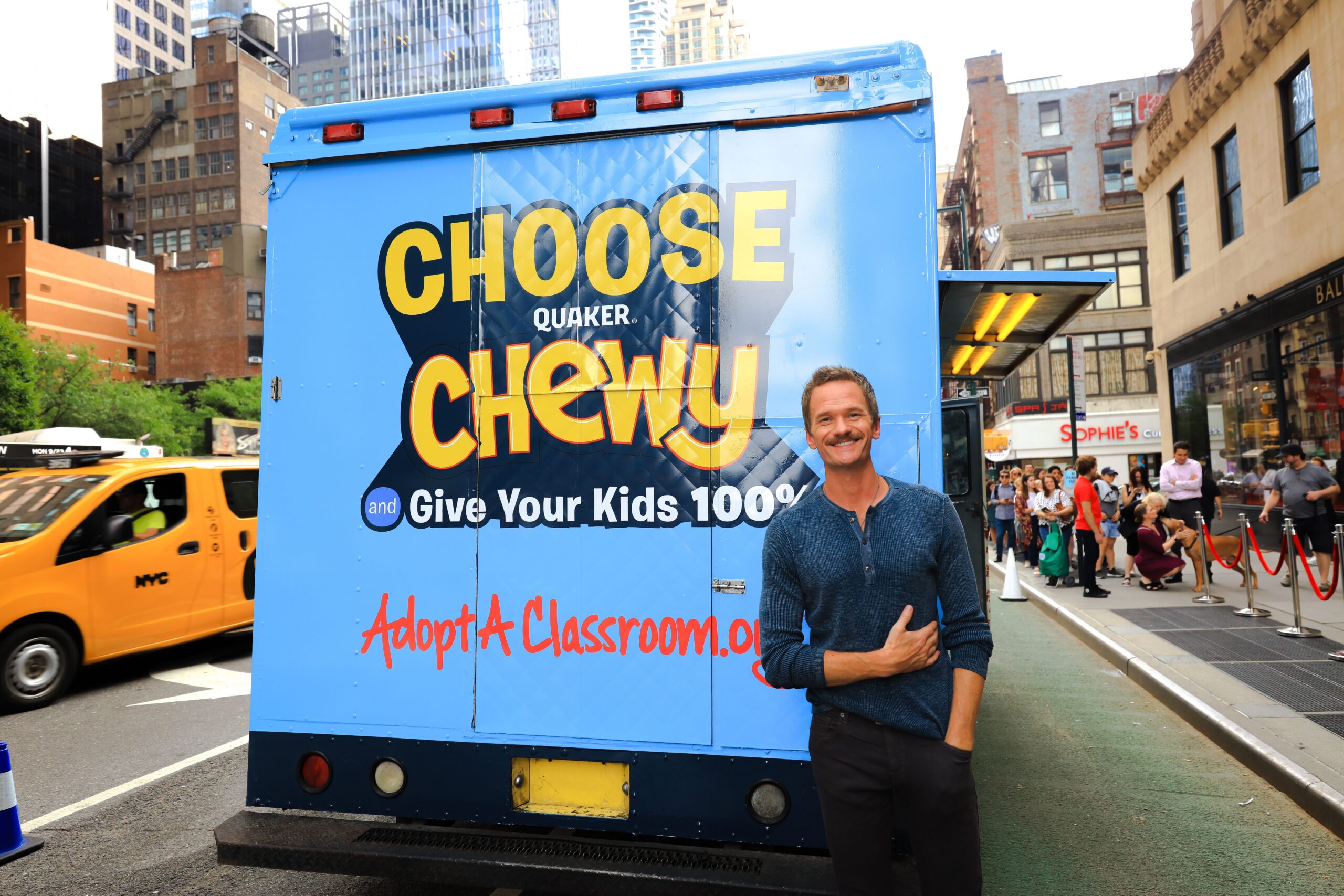 Man in front of blue food truck with Quaker Chewy sign, city street and yellow NYC taxi behind.