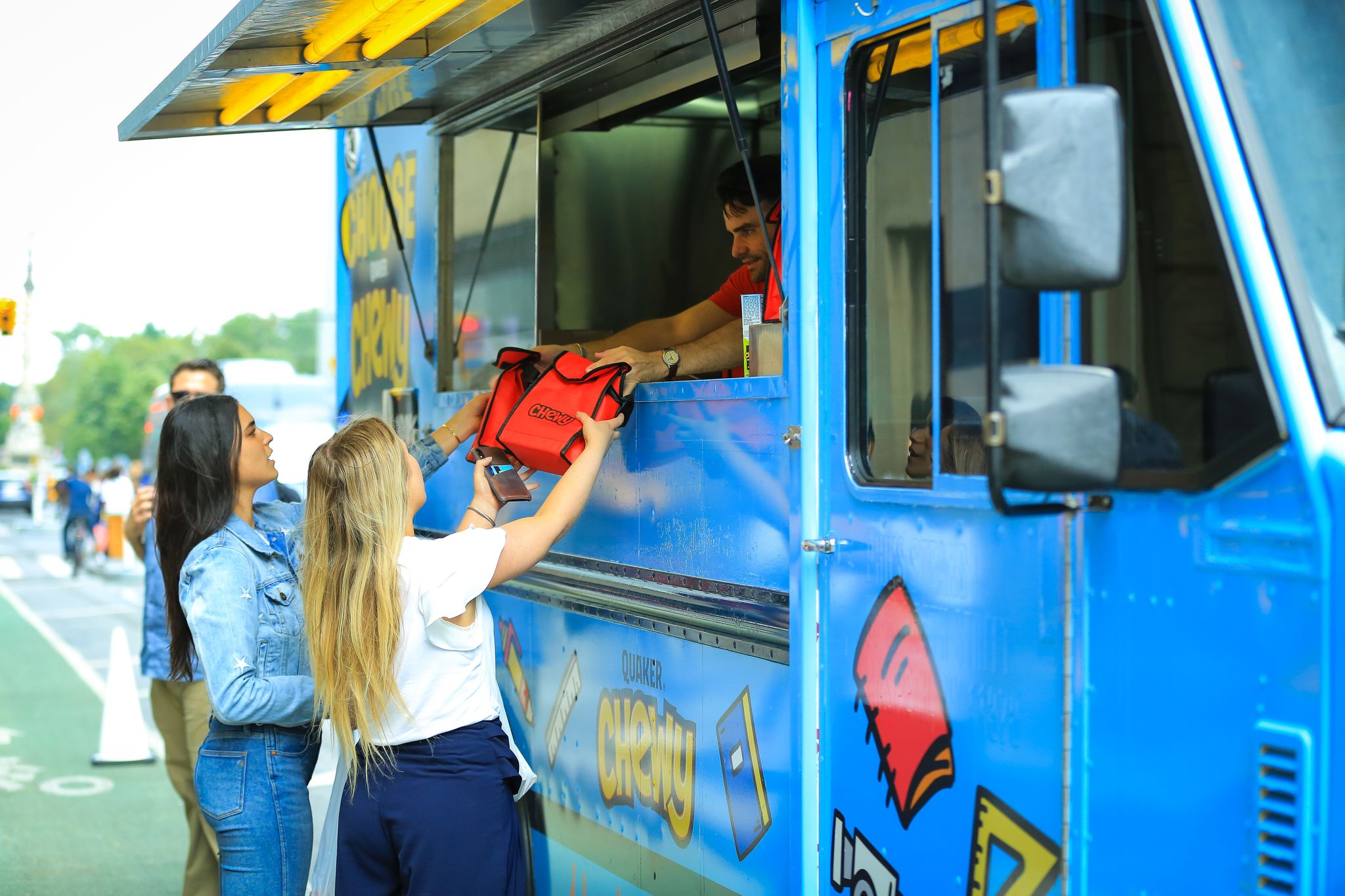 Two women at a blue food truck receive a red insulated food bag from a worker outside.