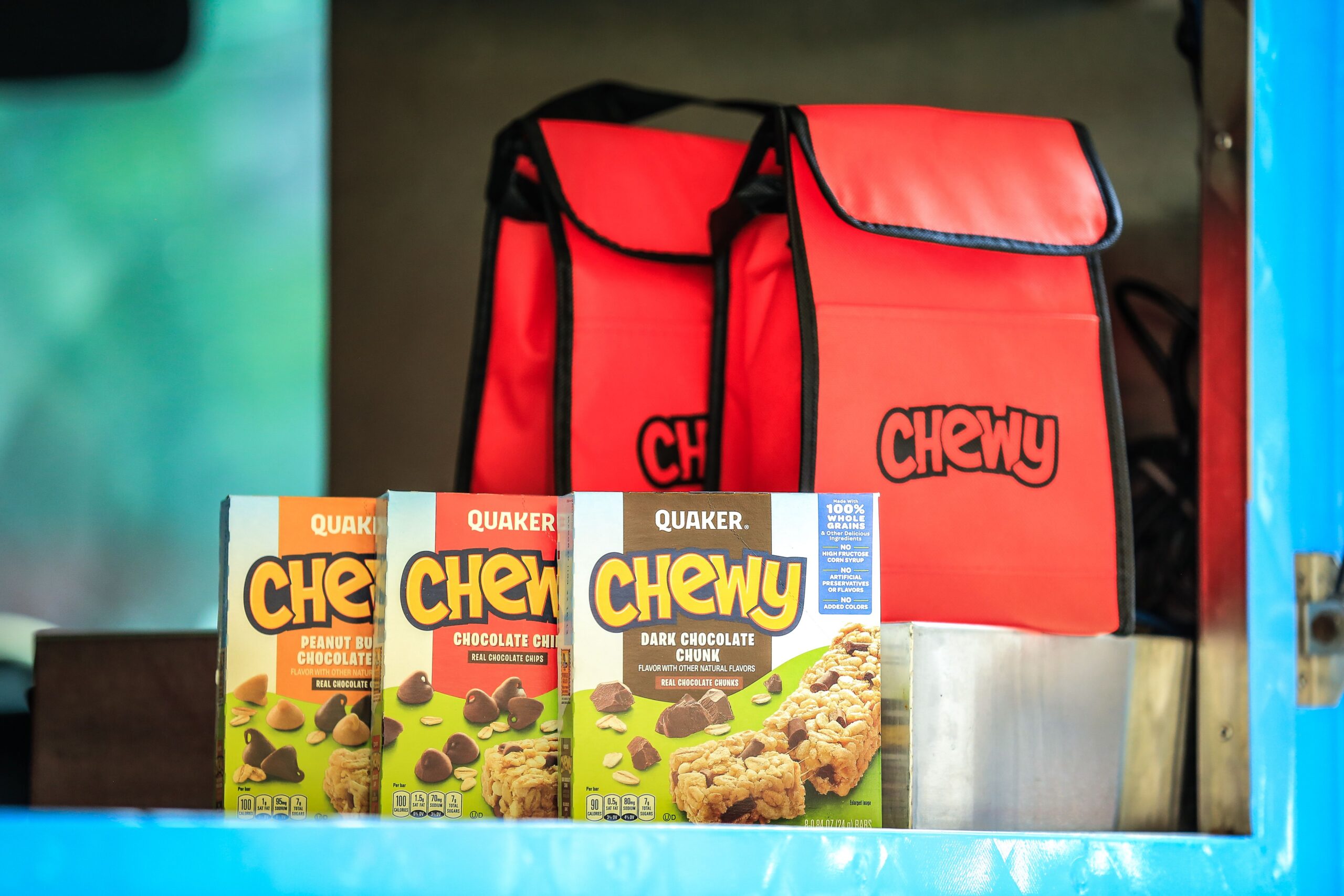 Three Quaker Chewy granola bar boxes in front of two red bags labeled Chewy.