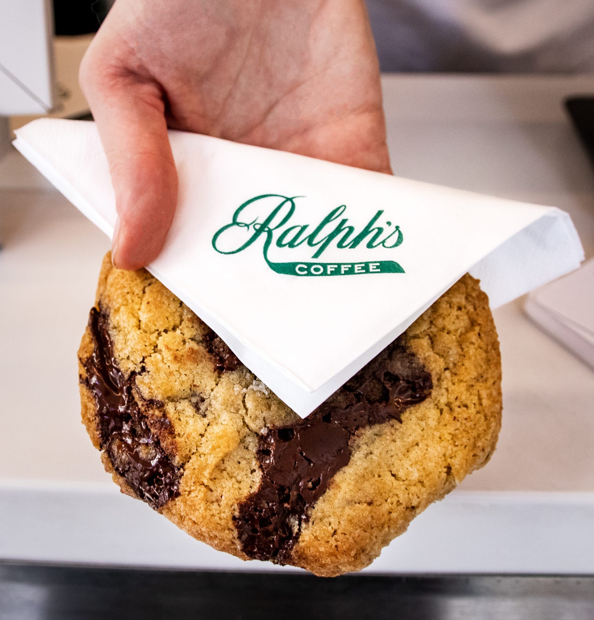 Hand holding a chocolate chip cookie in a white napkin labeled Ralph's Coffee.