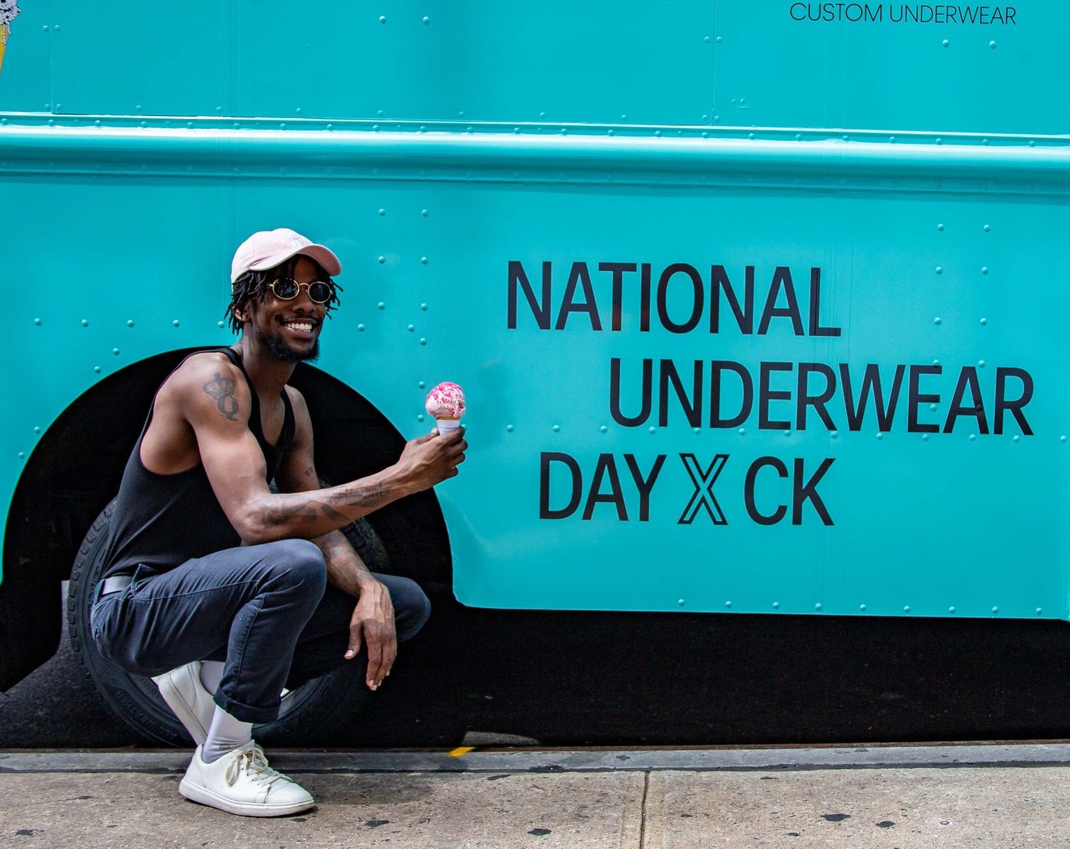 Man in sunglasses, pink cap, and black tank top smiles holding ice cream by a turquoise CK truck.