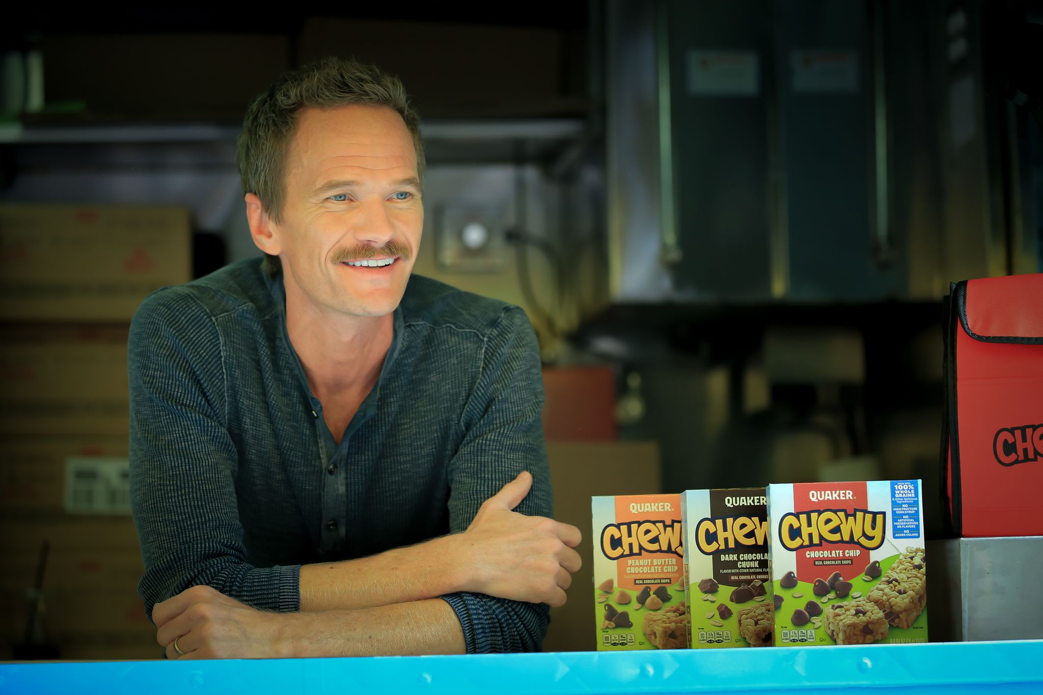 Man with mustache leans on counter by three Quaker Chewy granola bar boxes in a kitchen or truck.