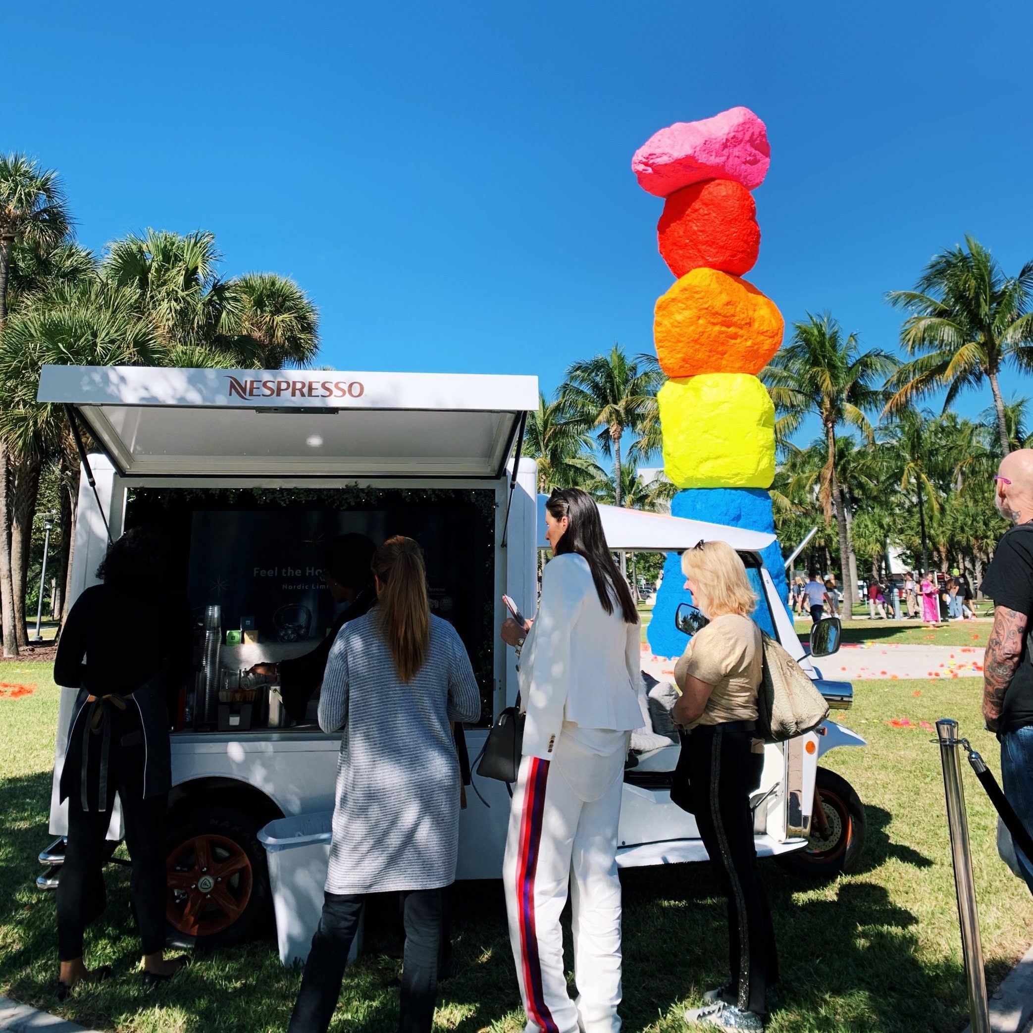 People line up at a Nespresso truck in a park with palm trees and stacked colorful boulders nearby.