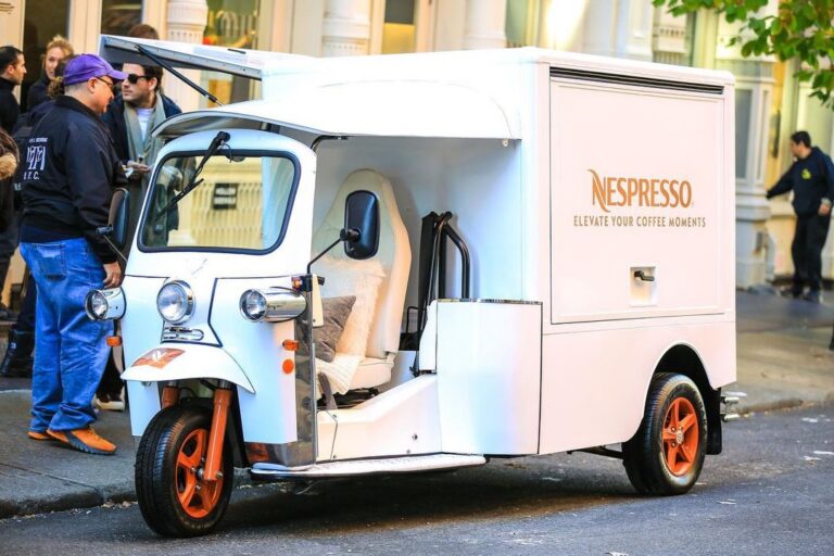 Nespresso three-wheeled vehicle with orange wheels parked on street; people and buildings nearby.