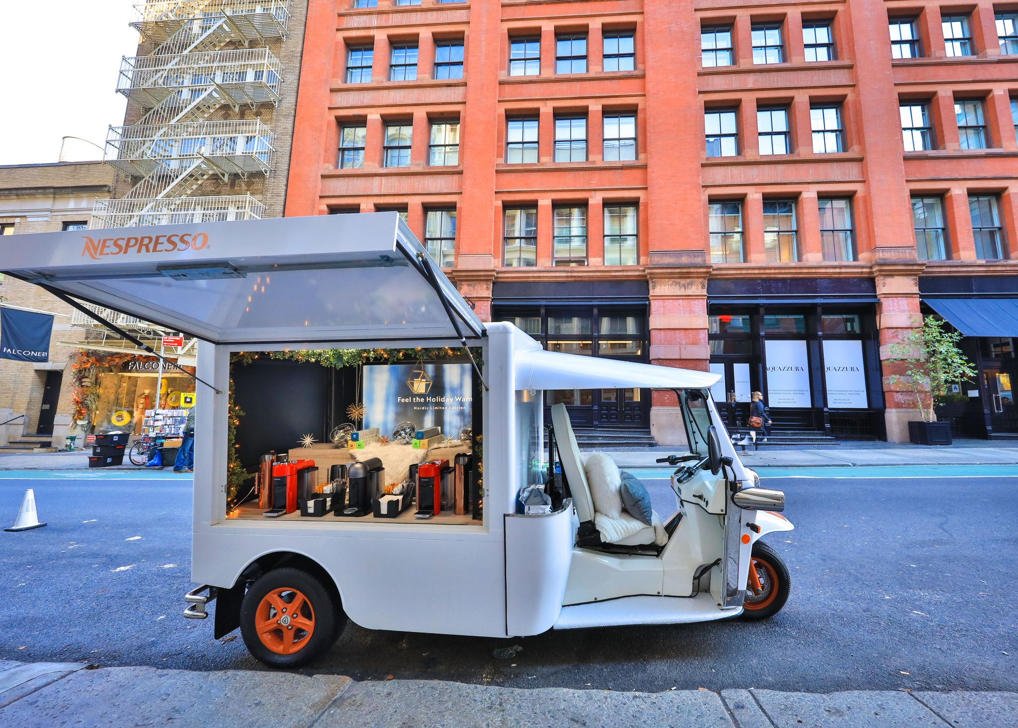A white Nespresso tuk-tuk truck displays coffee machines on a city street by a red brick building.