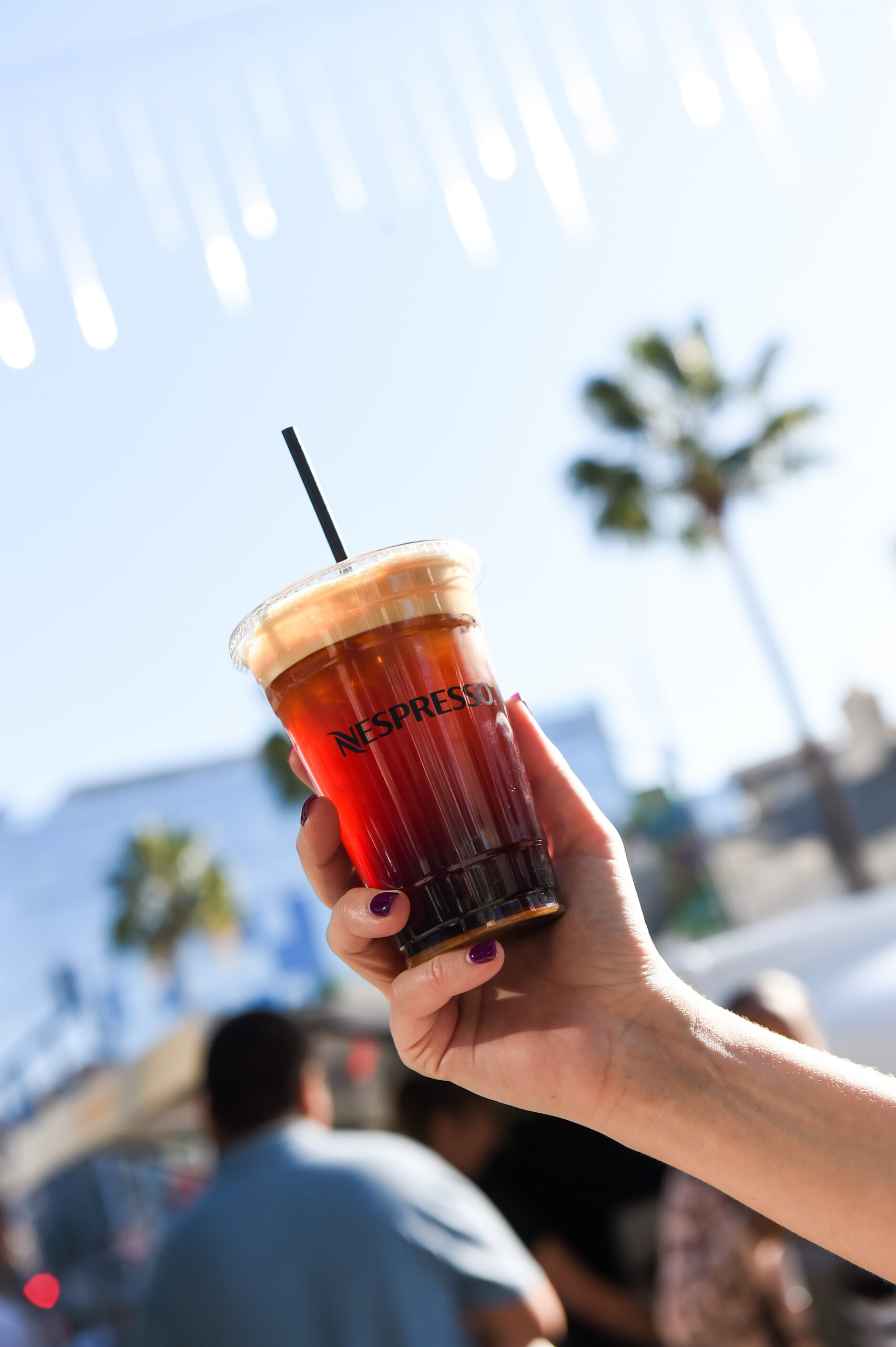 Hand with purple nails holds iced coffee in Nespresso cup; palm trees and people in background.