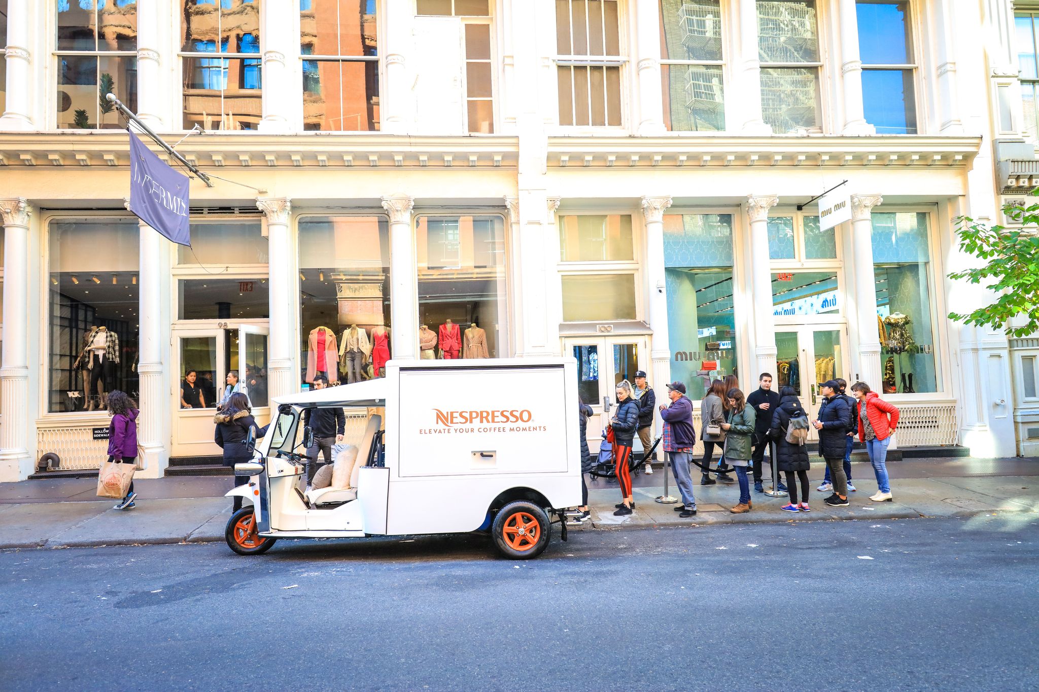 Nespresso mobile showroom parked on city street; people line up outside clothing store.