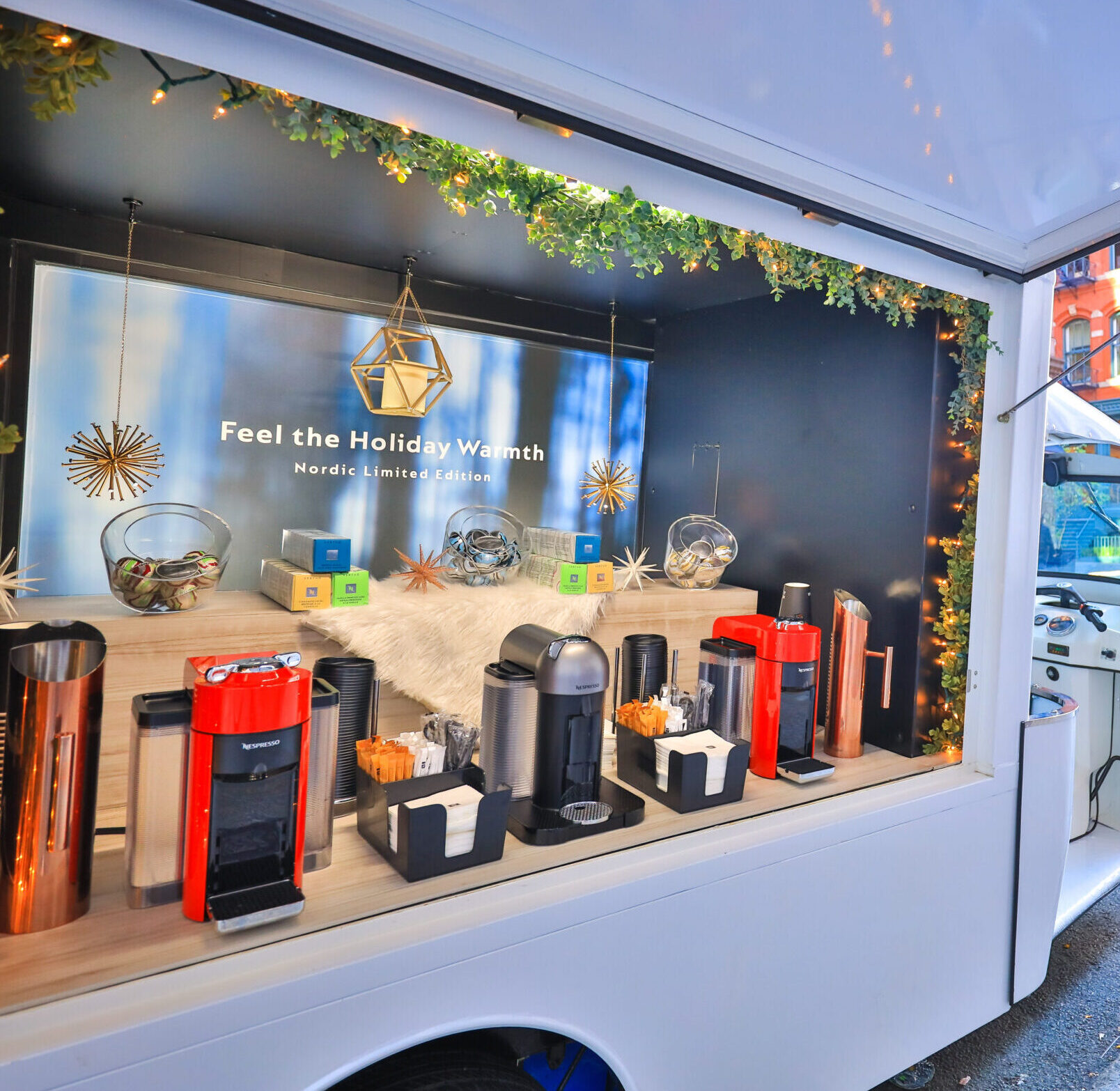Coffee truck display with coffee machines, cups, holiday decor, lights, greenery, and a promo sign.