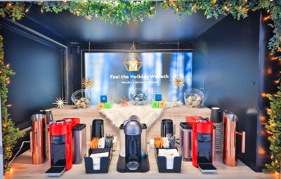 Coffee station with three machines, cups, stirrers, sign, greenery, and string lights.