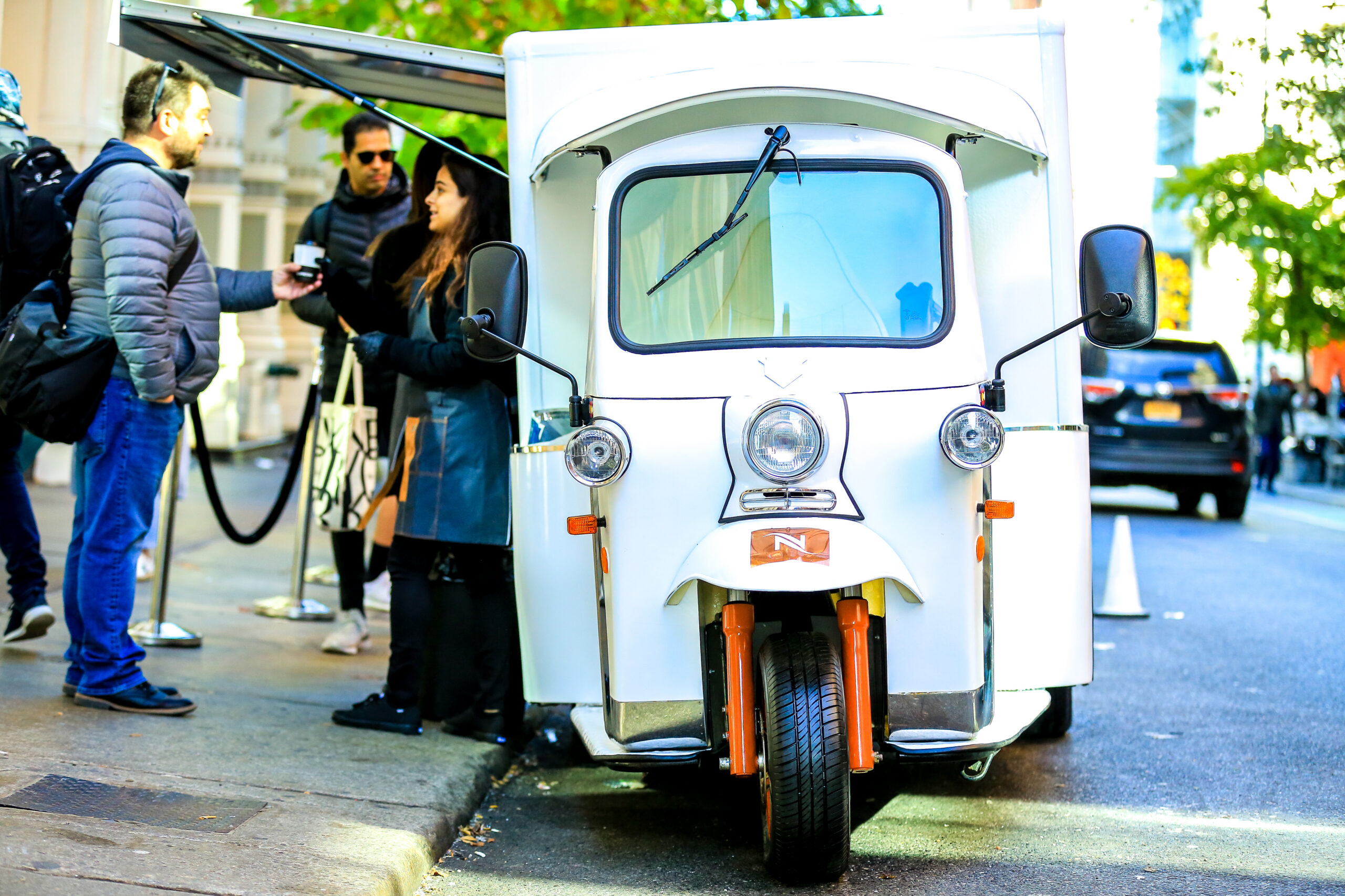 People wait as a white three-wheeled vehicle serves coffee at the curb; car and trees behind.
