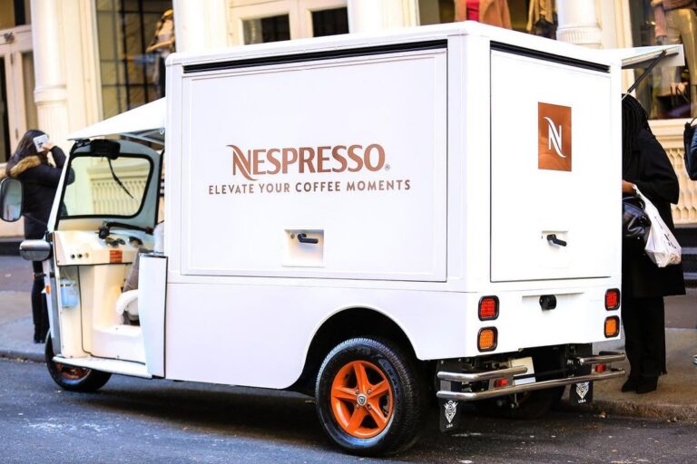 Nespresso mobile showroom van with orange wheels parked on city street; people stand nearby.