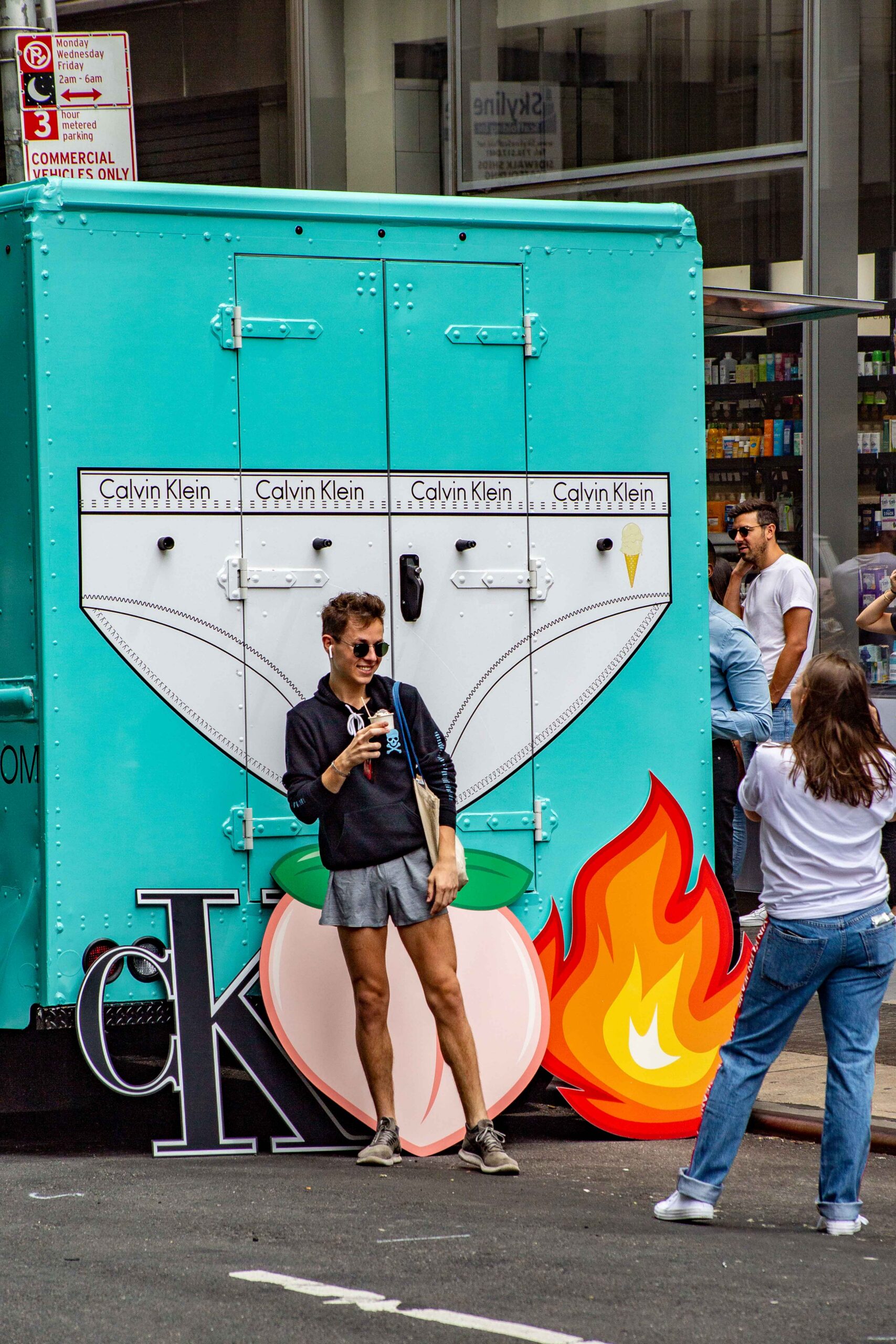 Person in sunglasses poses by Calvin Klein truck as another takes their photo on a city street.