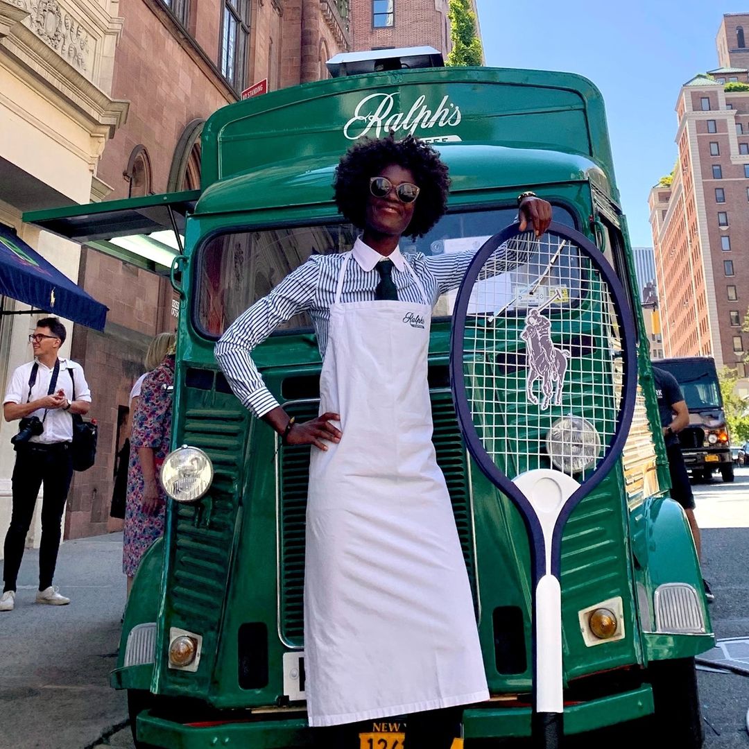 Person in striped shirt and apron holds large tennis racket by green Ralph's truck on city street.