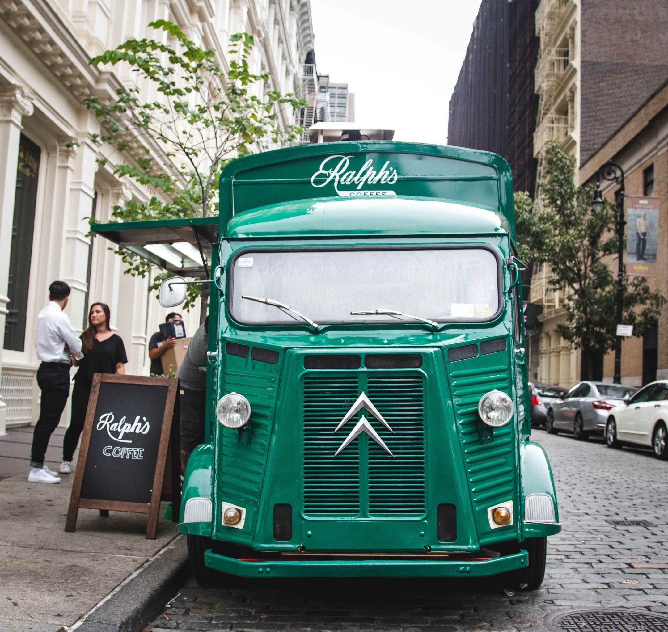 Green Ralph's Coffee truck on city street; two people by a sign, tall buildings in background.