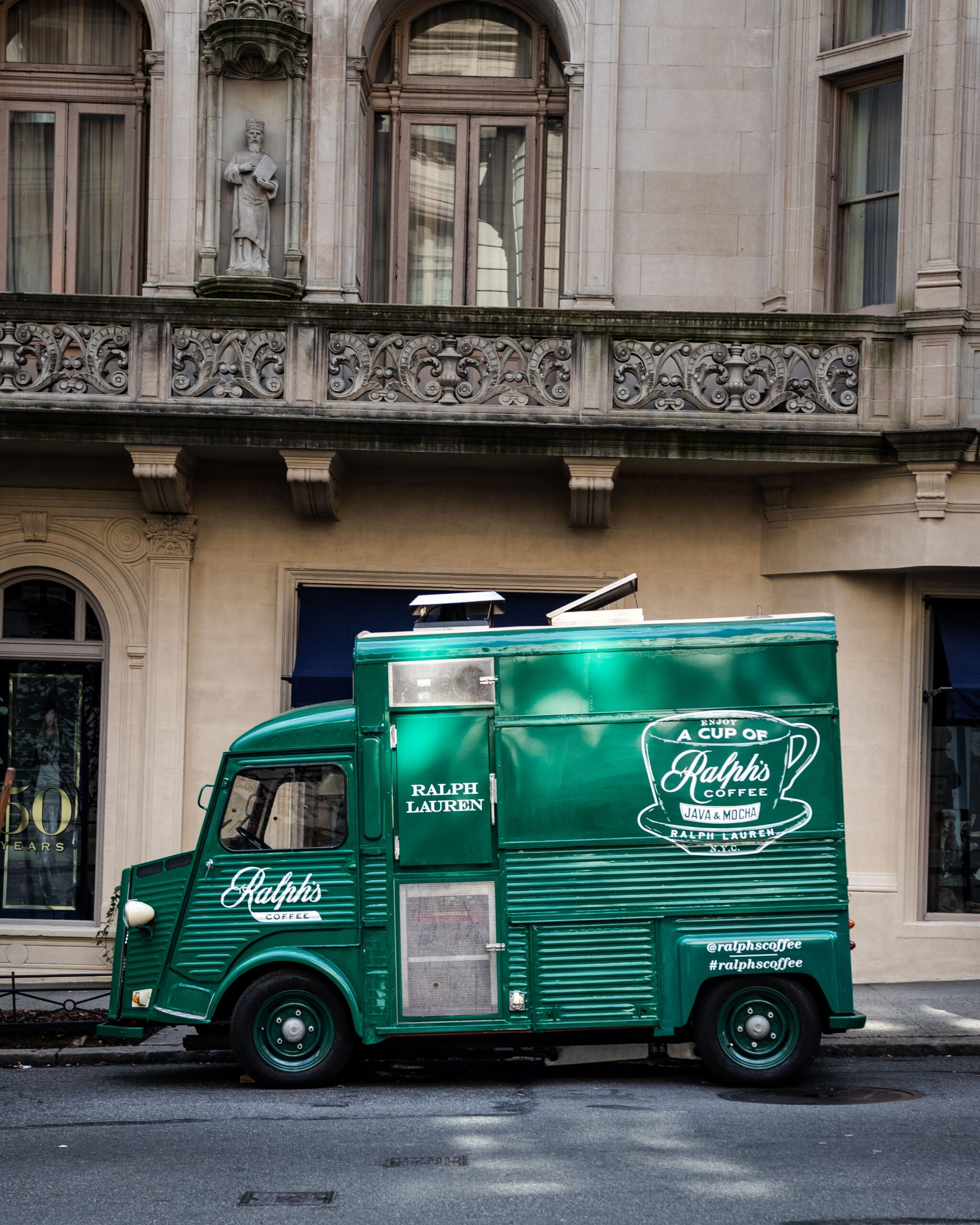 A green Ralph’s Coffee food truck parked on a city street by a stone building with balconies.