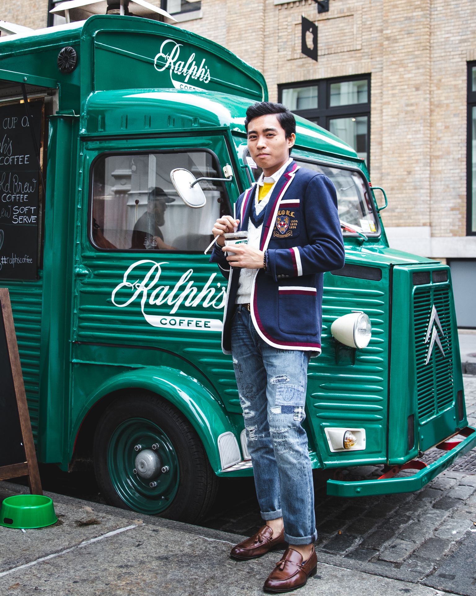 Man with coffee stands by a green Ralph’s Coffee truck on a cobblestone street near brick buildings.