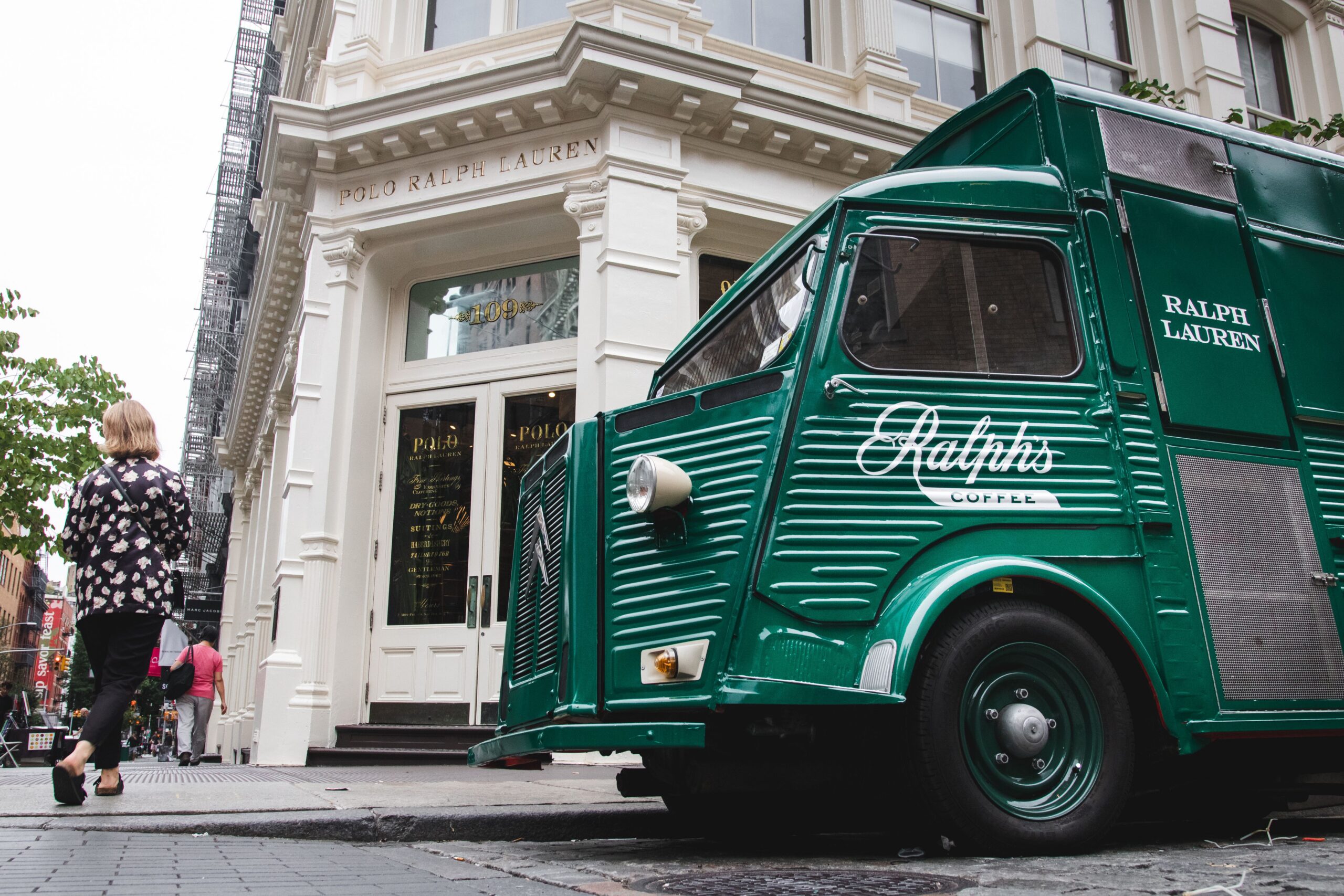 Green Ralph’s Coffee truck parked by Polo Ralph Lauren store as woman in patterned shirt walks past.