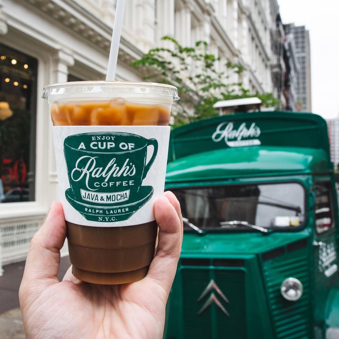 Hand holding an iced coffee in front of a green Ralph’s Coffee truck on a busy city street.