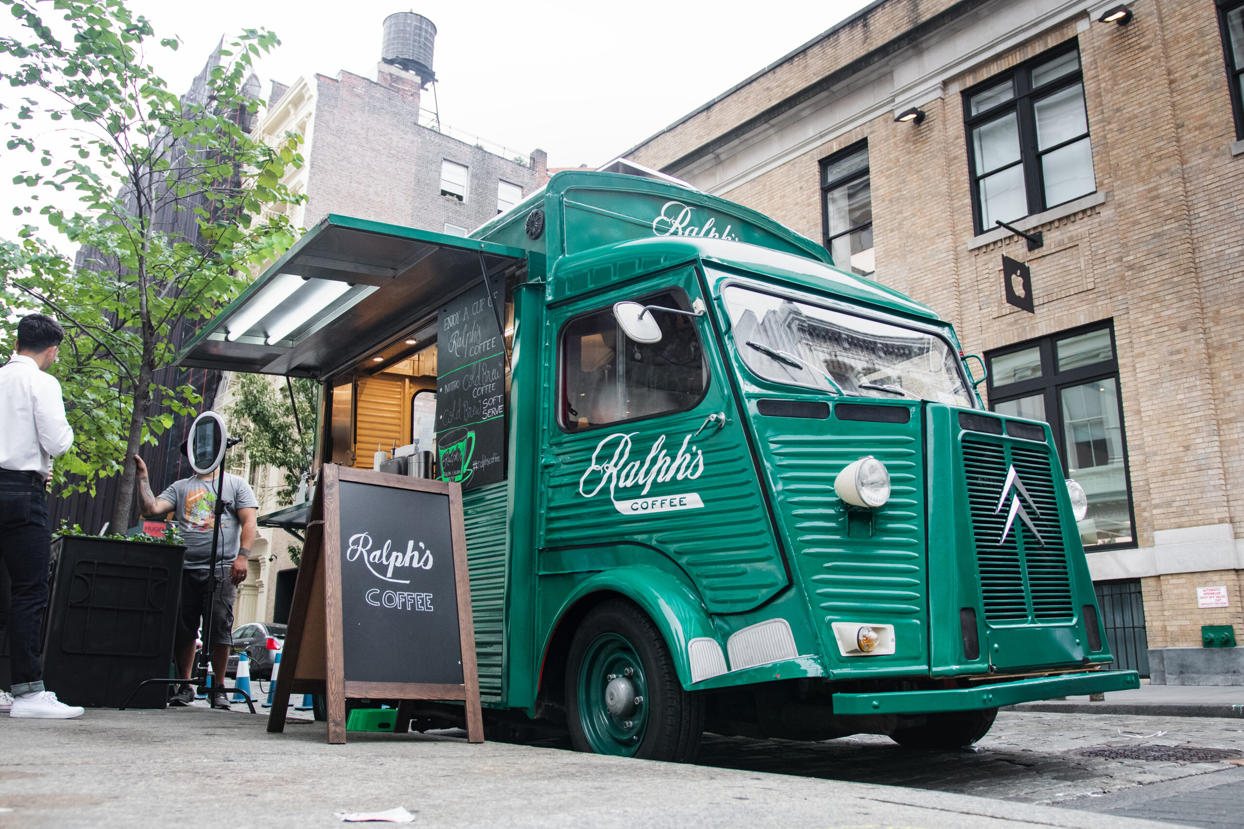 Green "Ralph's Coffee" truck on city street with chalkboard menu and people nearby.