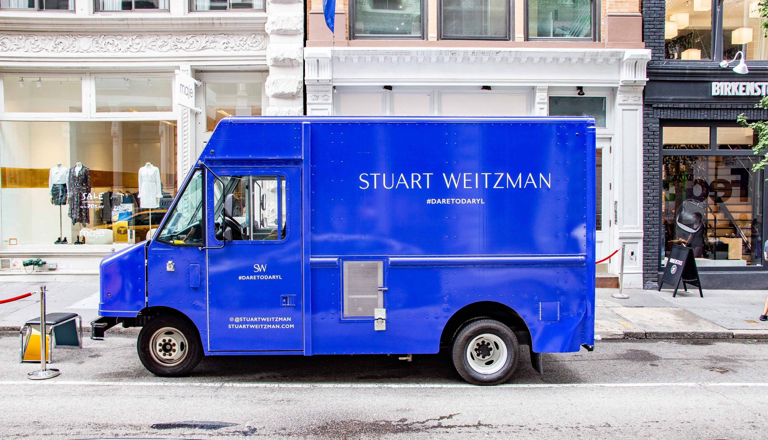 Blue STUART WEITZMAN food truck parked on city street in front of clothing store windows.