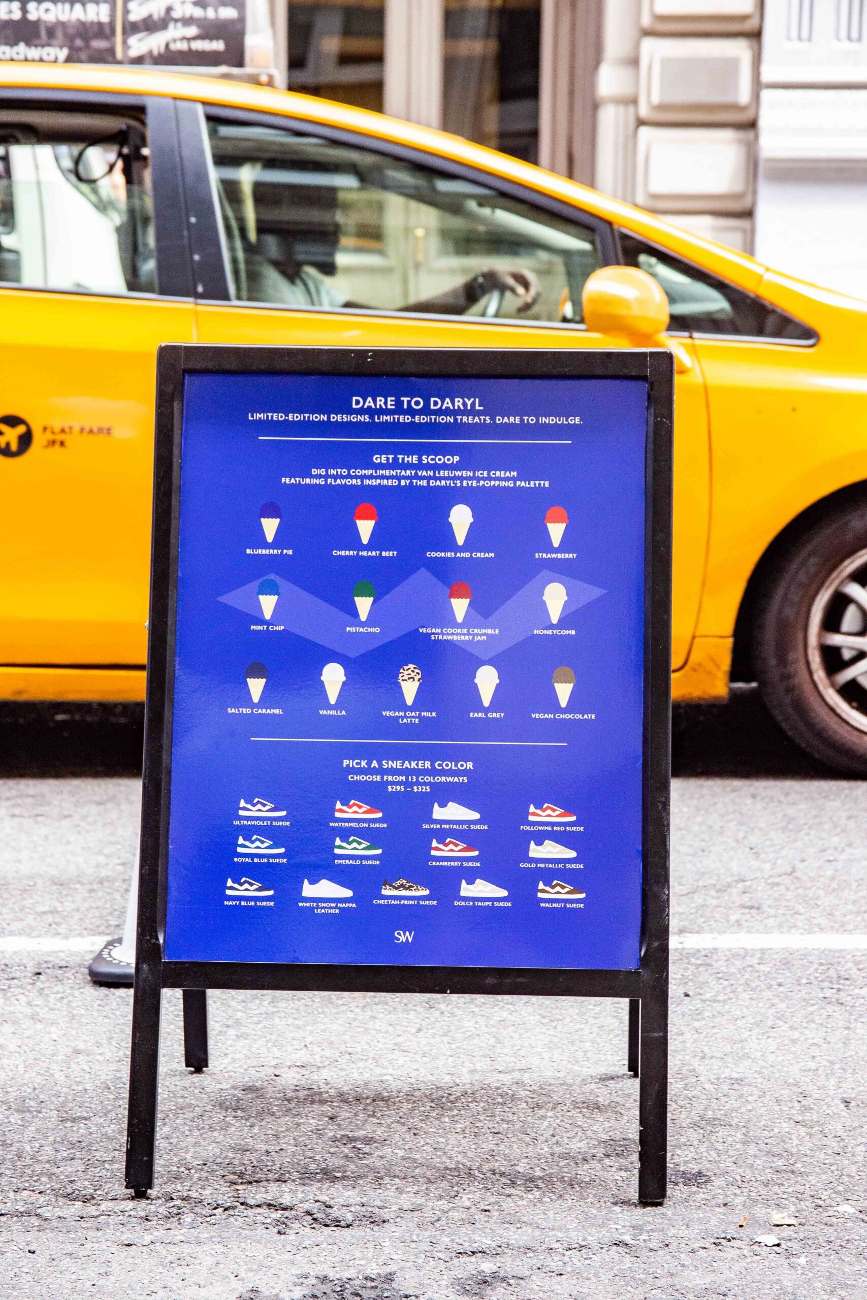Blue sidewalk sign by yellow taxi shows ice cream and sneakers, offering flavor and color choices.