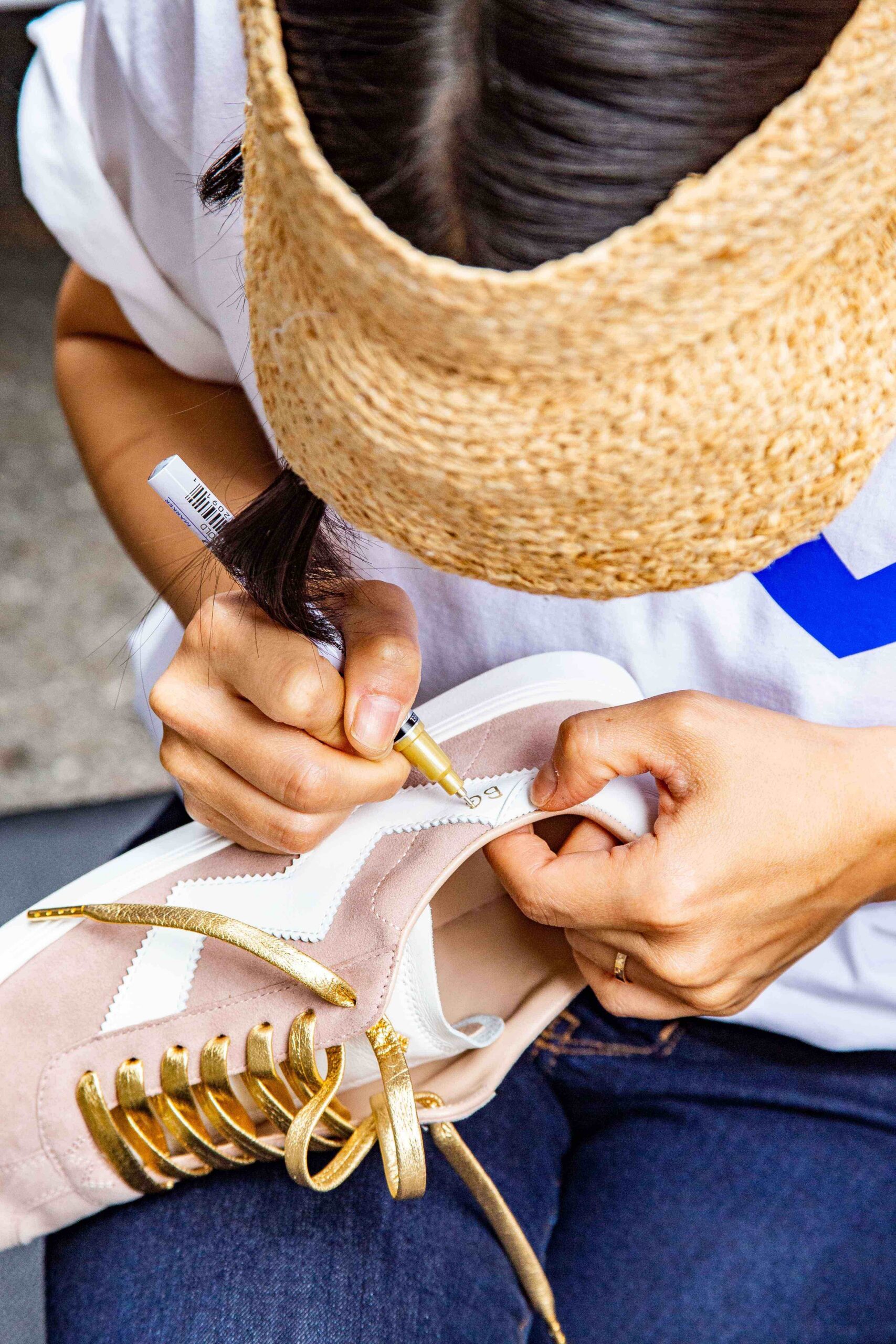 Person in a straw visor decorates a beige and white sneaker with gold laces using a gold marker.