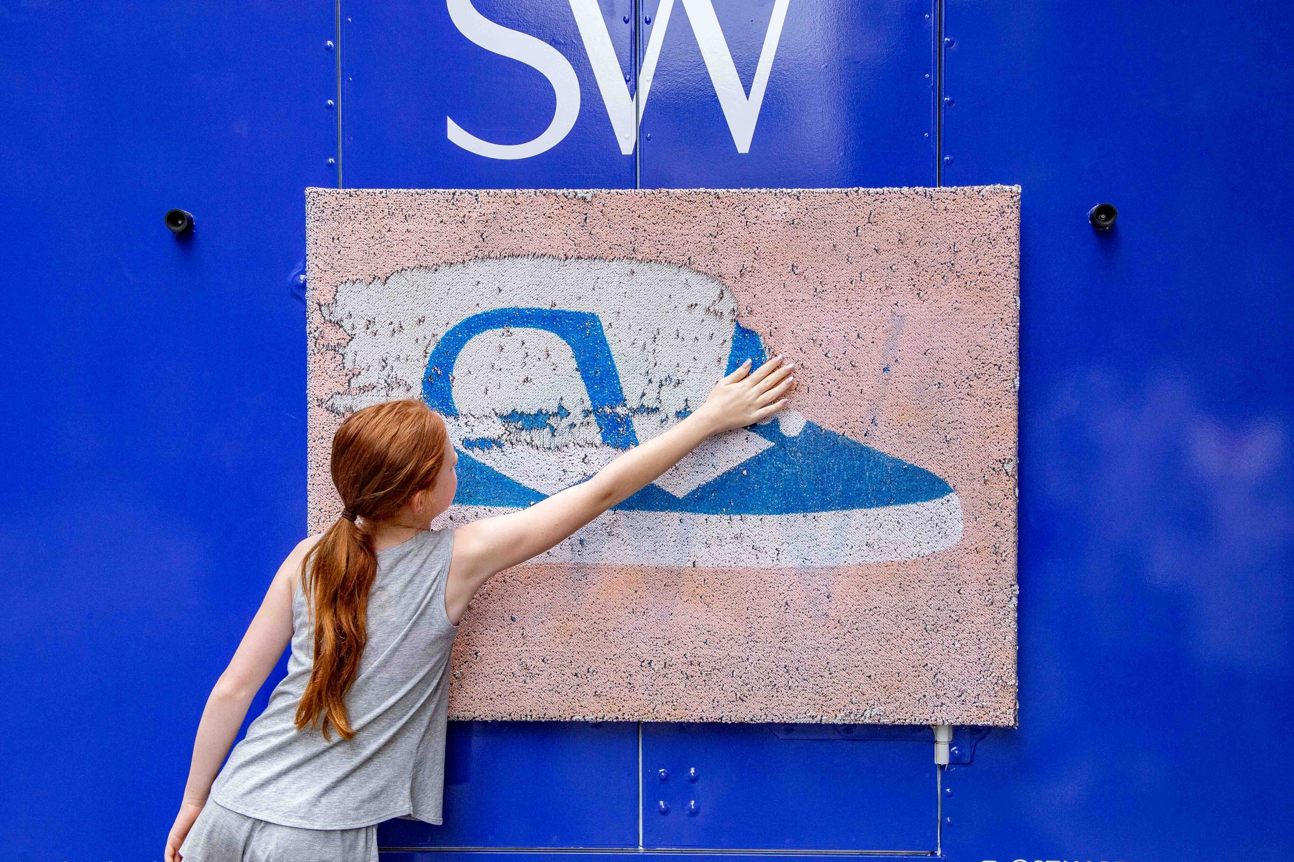 Girl with red ponytail touches textured blue and white sneaker mural under large SW letters.