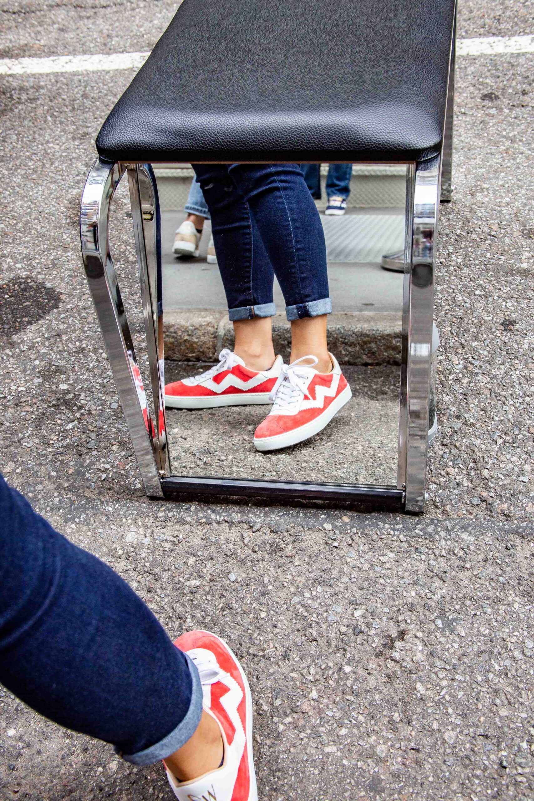 Person in red and white sneakers and blue jeans stands on street, legs reflected in mirror.