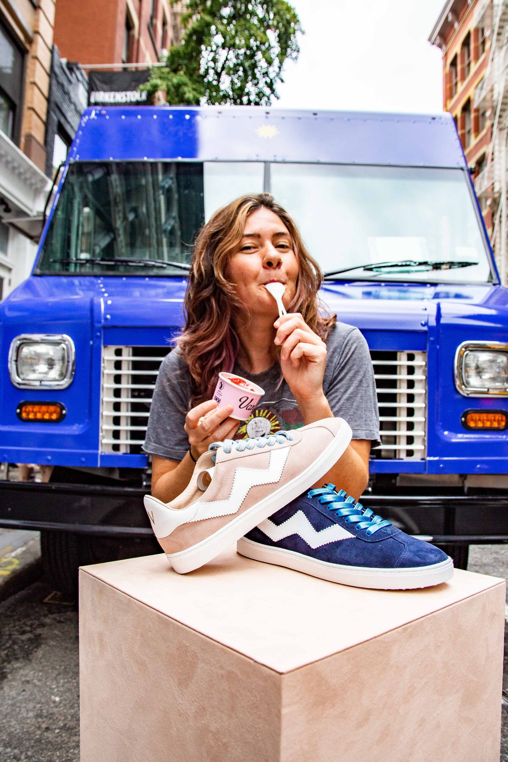 Woman eating ice cream near a blue truck with two pairs of sneakers on a light pedestal.